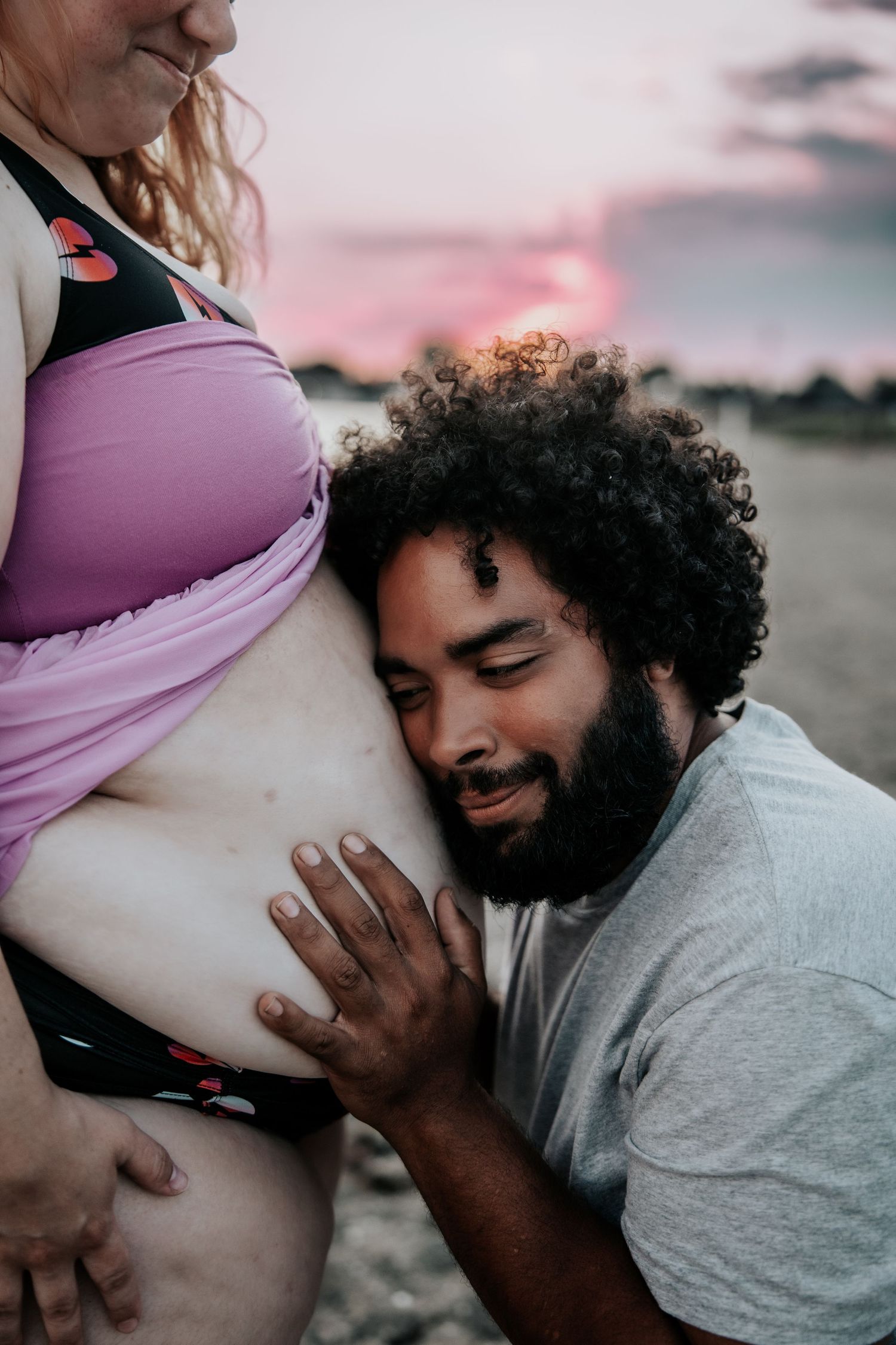 Man with curly hair and beard tenderly listens to pregnant womans belly at sunset beach.