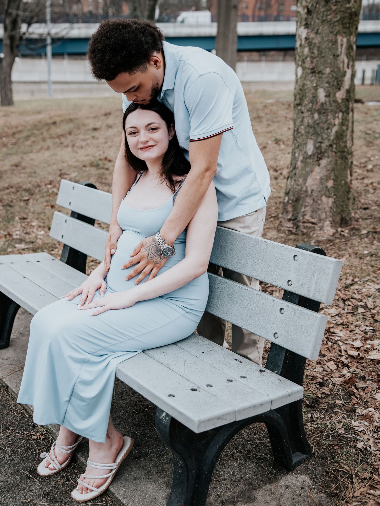 Couple sharing a tender moment on a park bench, man embracing pregnant woman in a light blue maternity dress.