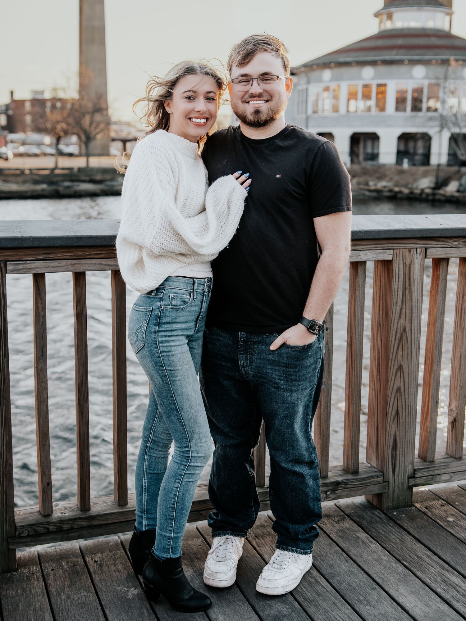 A smiling couple poses on a waterfront dock, woman in white sweater and jeans, man in black shirt and jeans.