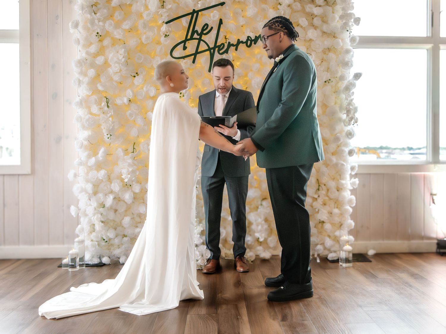 Couple exchanges vows at elegant wedding ceremony before floral backdrop, bride in white cape gown, groom in green suit.