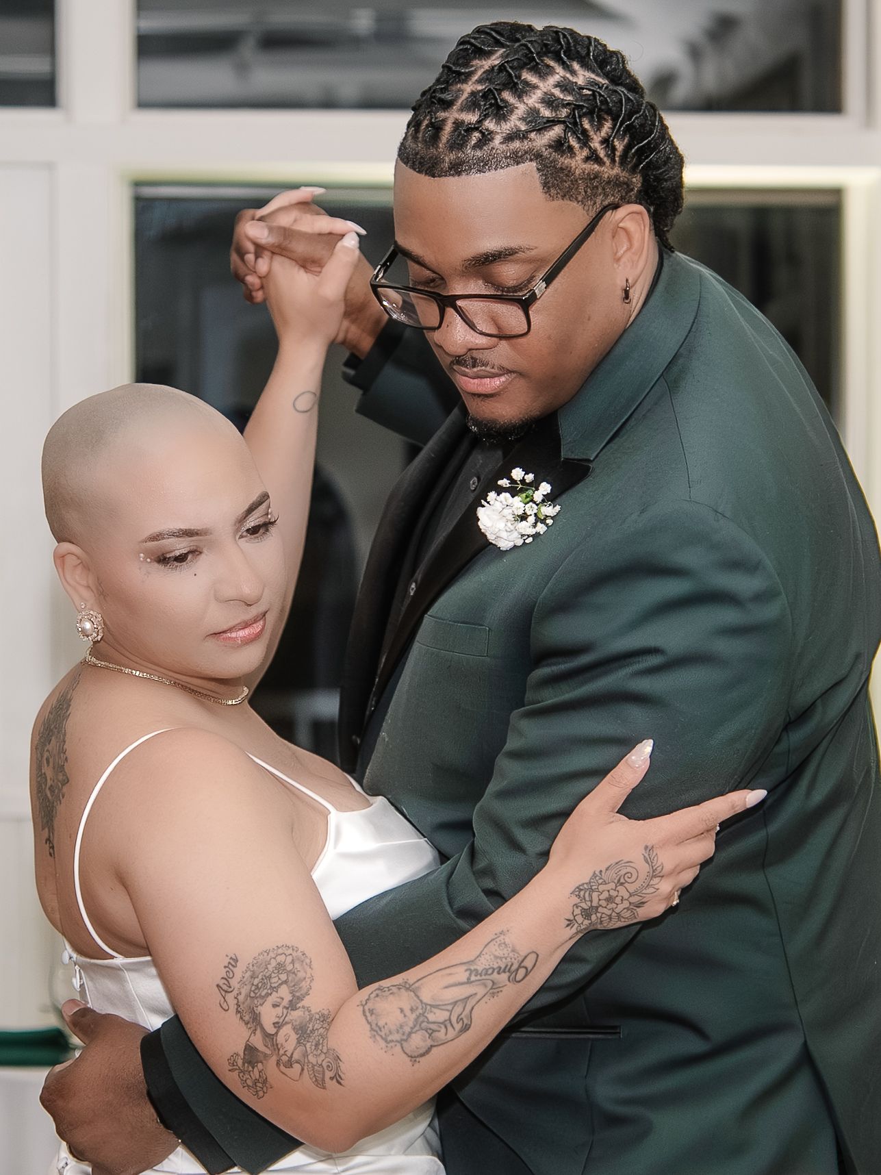 A couple shares a romantic dance, woman with shaved head and tattoos in white dress, man in green tuxedo.