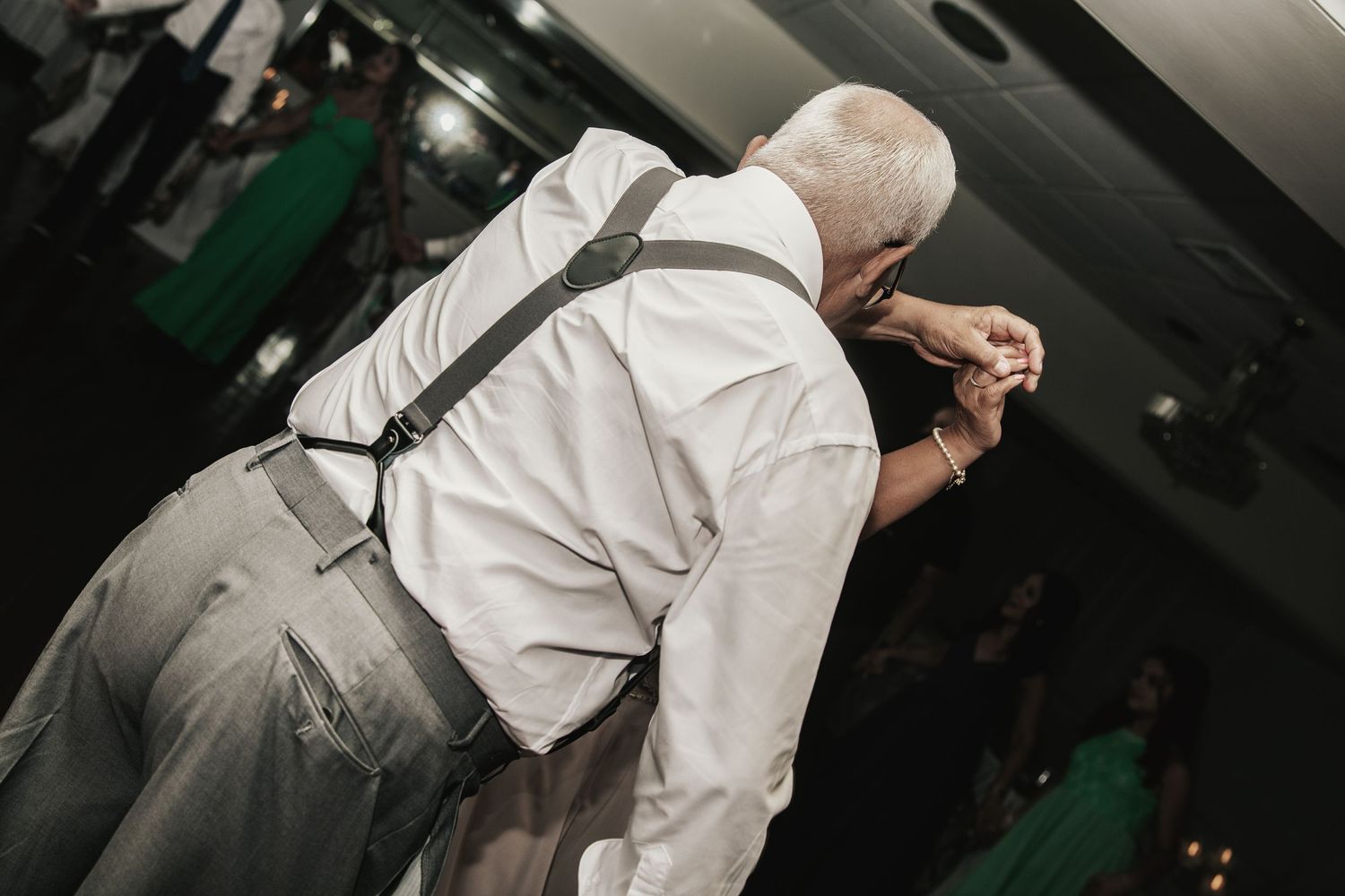 Elderly man in white shirt and suspenders dancing at a formal event or wedding reception.
