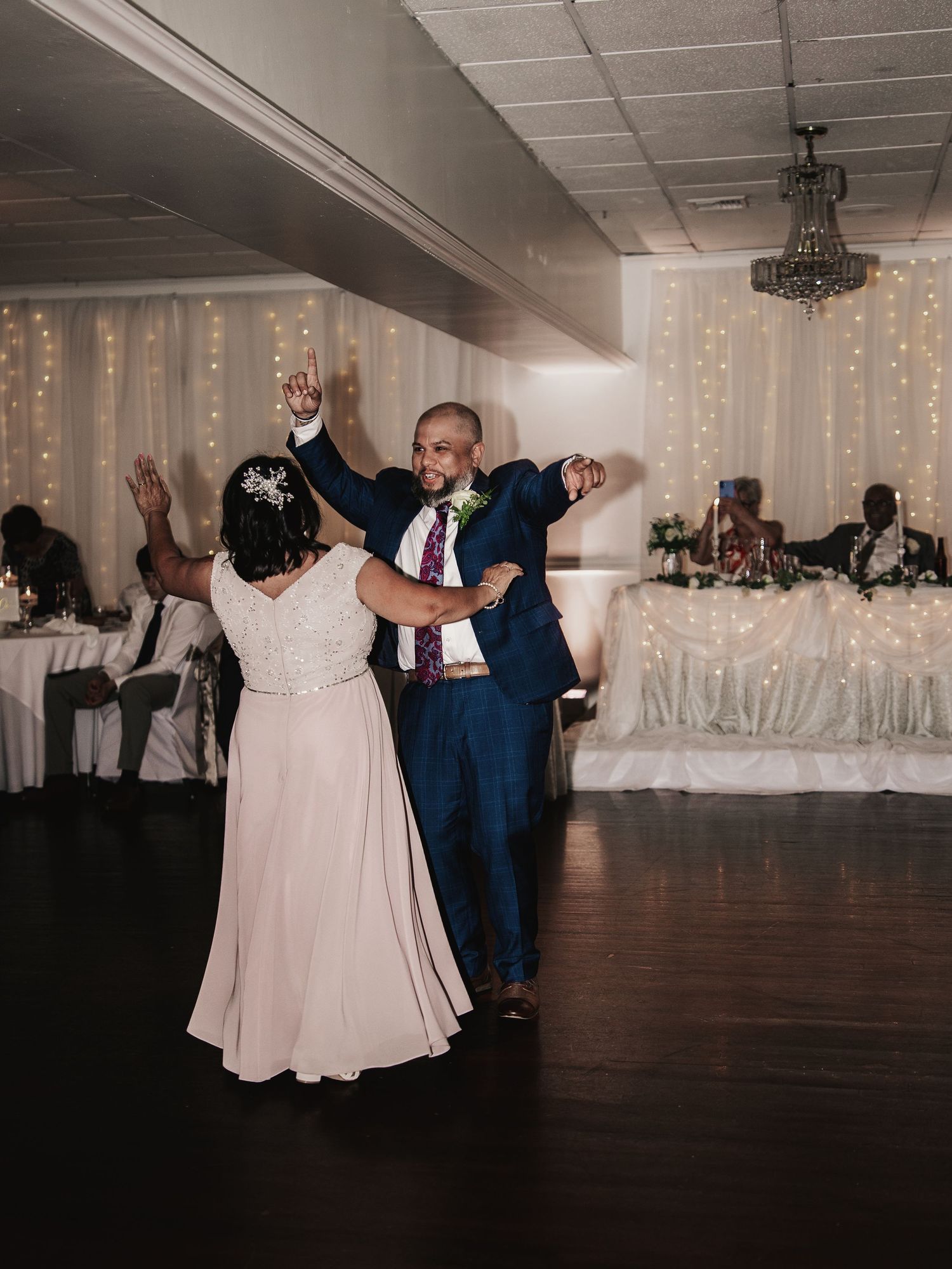 Couple sharing a joyful first dance at wedding reception with fairy lights and chandelier backdrop.