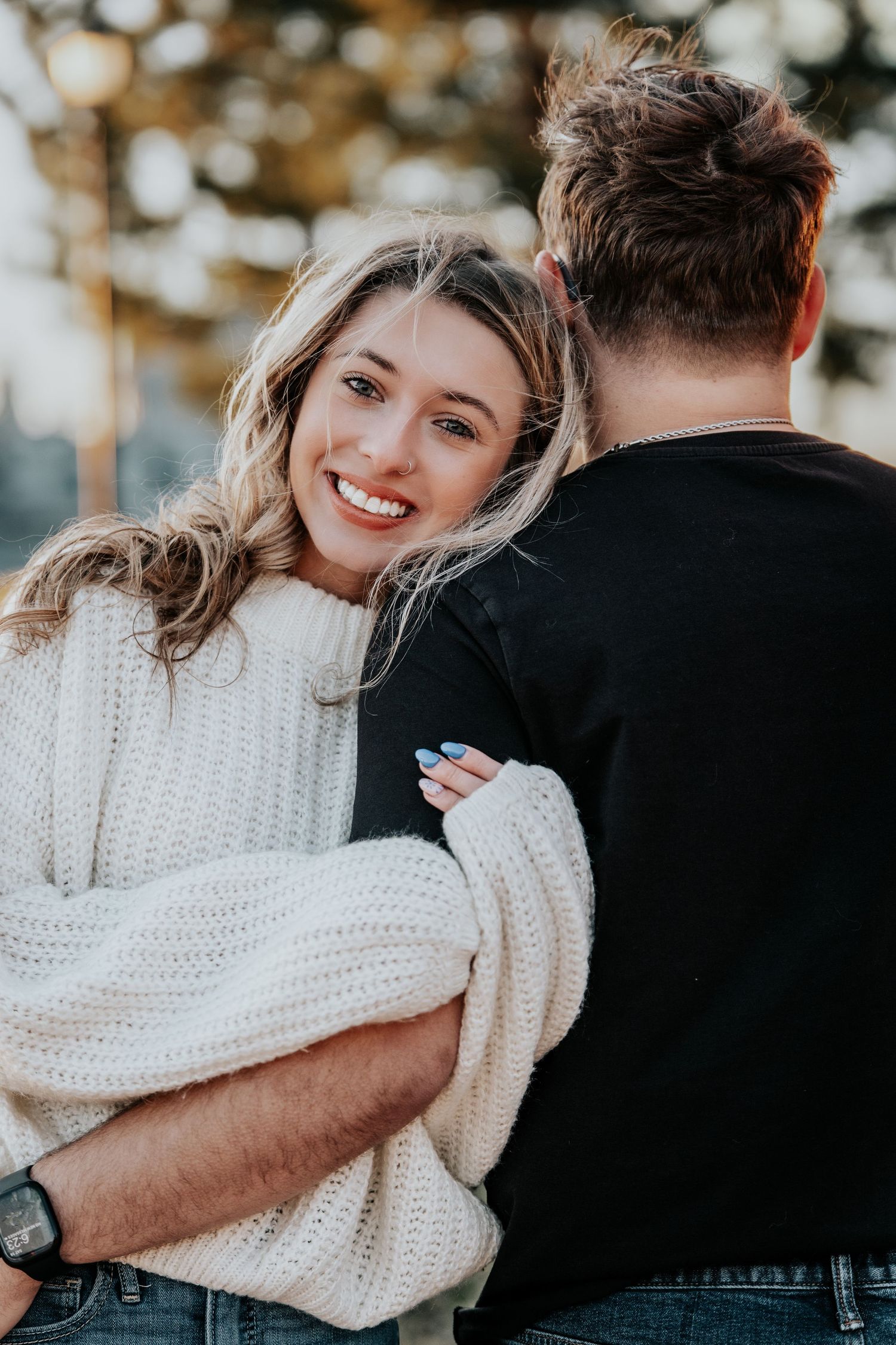 Smiling blonde woman in white knit sweater hugging man in black shirt from behind outdoors.