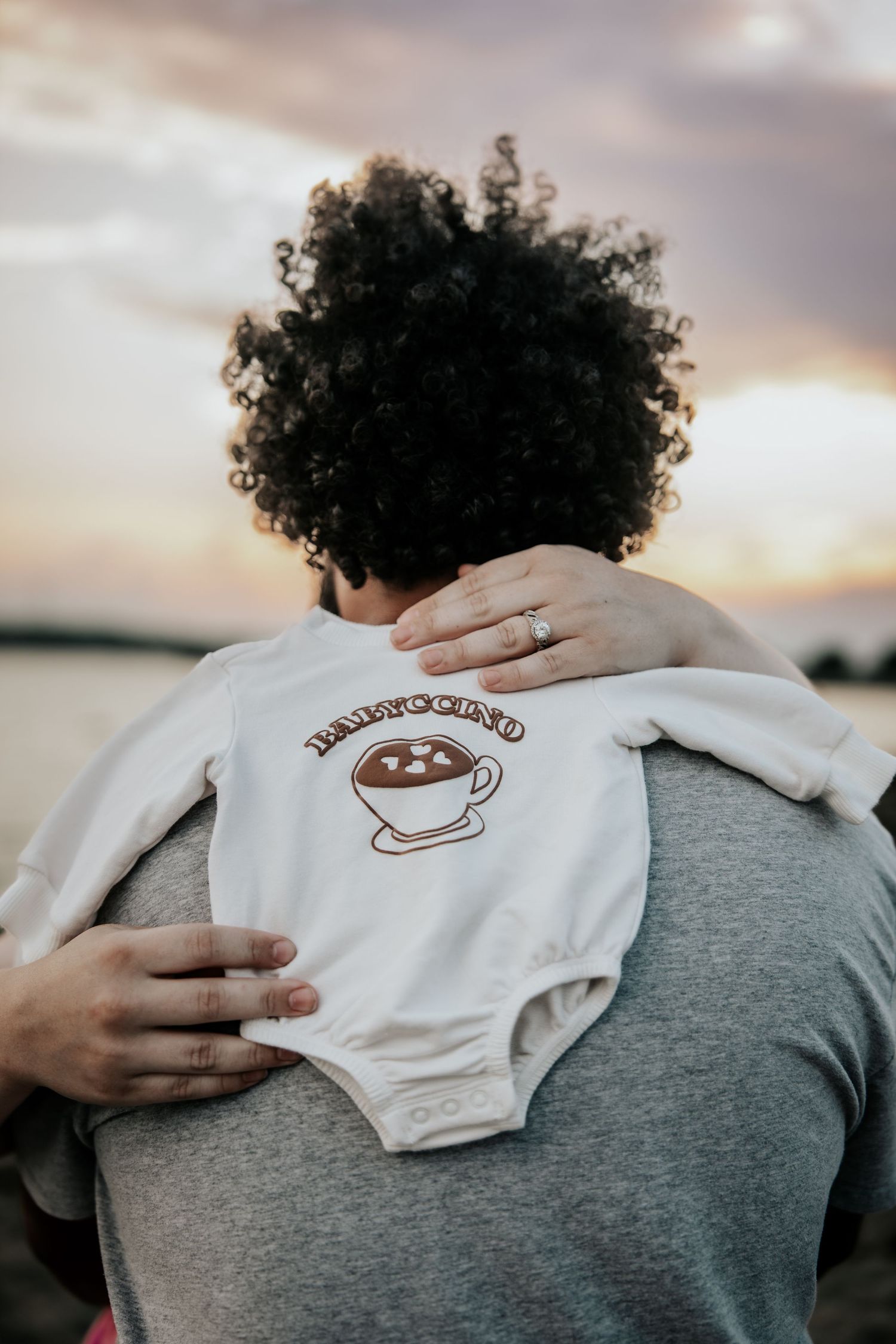 Parent holding baby in white 'Babyccino' onesie with coffee cup graphic at sunset near water.