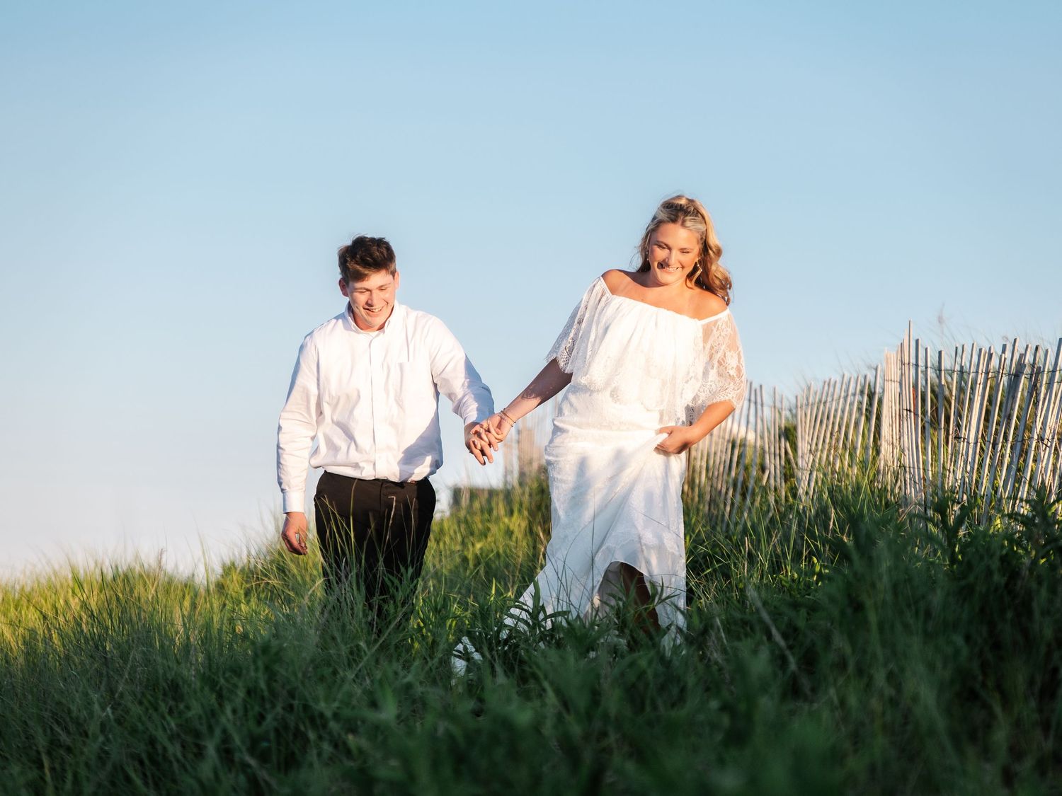 Joyful couple holding hands and running through green grass near a wooden fence on a sunny day.