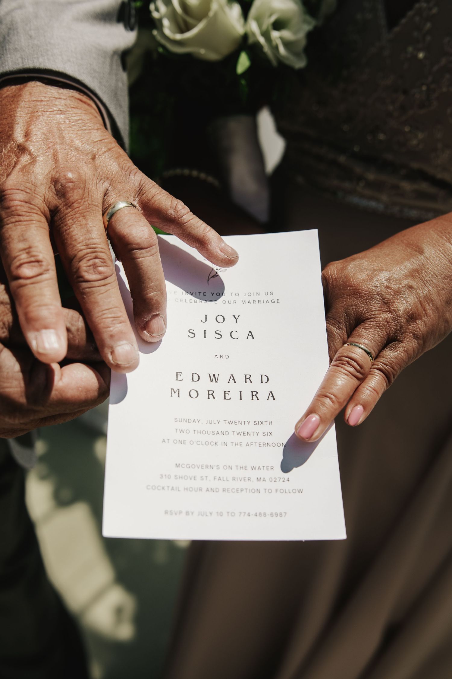Elderly hands holding a wedding invitation for Joy Sisca and Edward Moreira, one hand wearing a gold ring.