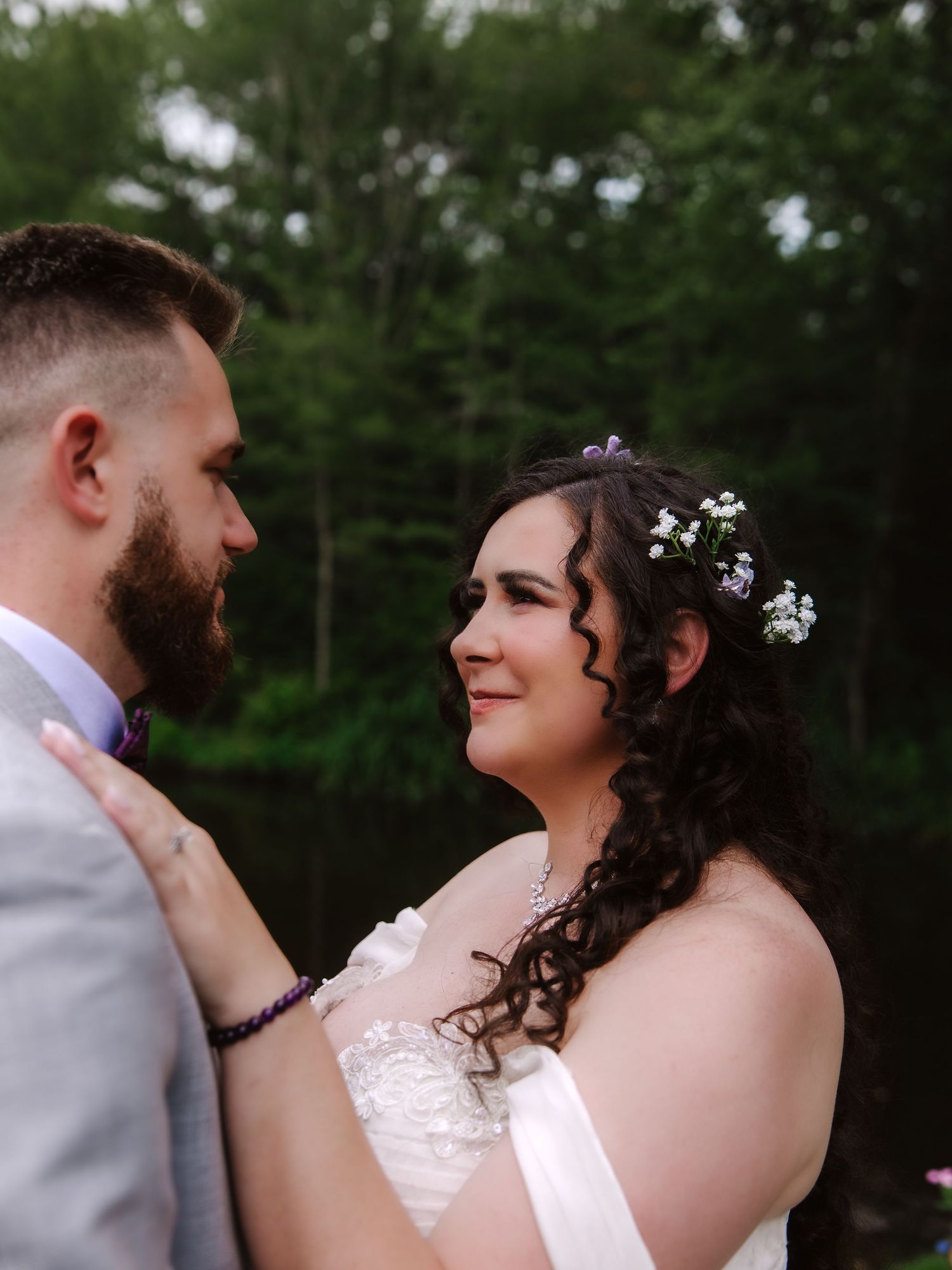 A bride and groom share a tender moment outdoors, she wearing a white gown with floral hair accessories, he in a gray suit.