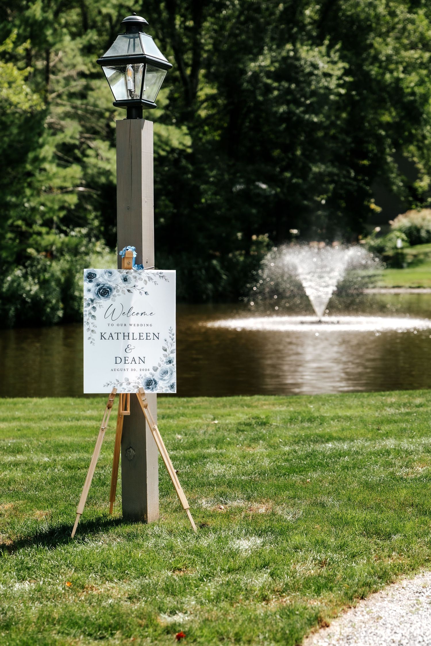 Wedding welcome sign for Kathleen and Dean on easel by lamp post with pond fountain backdrop.