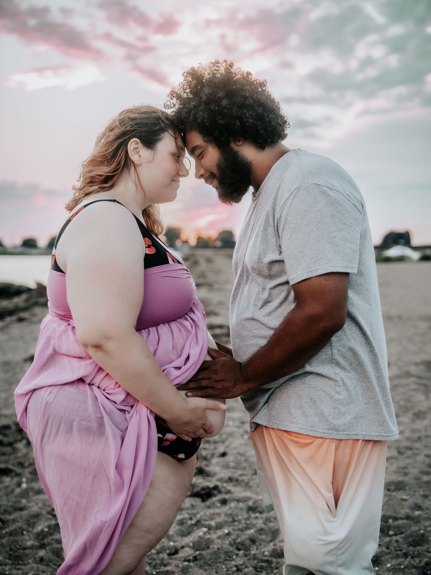 Pregnant couple holding hands on beach at sunset, woman in pink dress and man in gray shirt sharing tender moment.