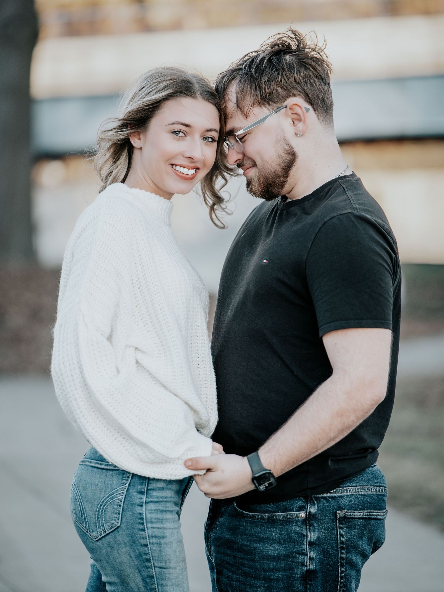 Smiling couple in white sweater and black tee holding hands outdoors during golden hour engagement photo.