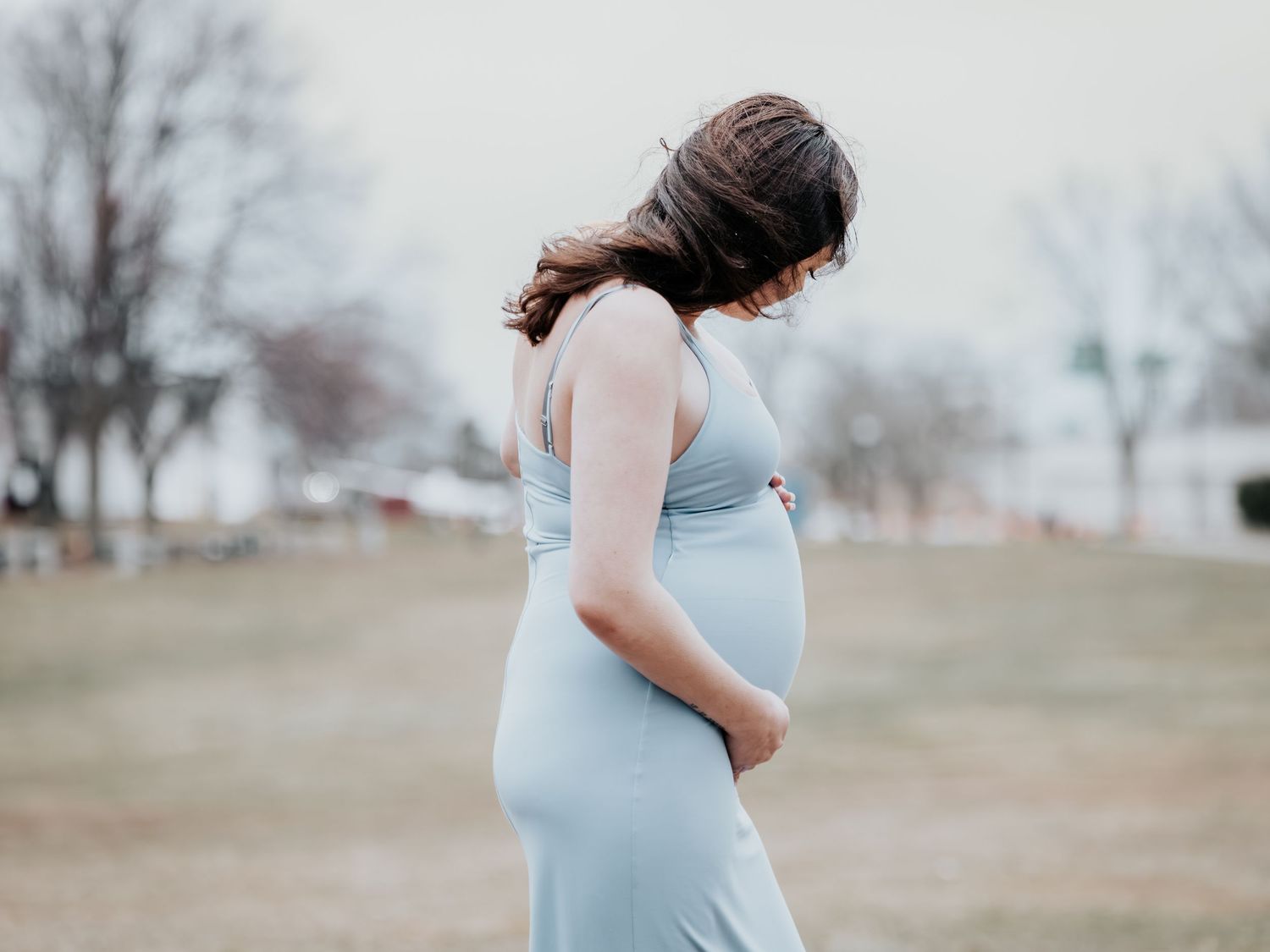 Pregnant woman in light blue dress cradling her baby bump outdoors in a park during a maternity photo shoot.