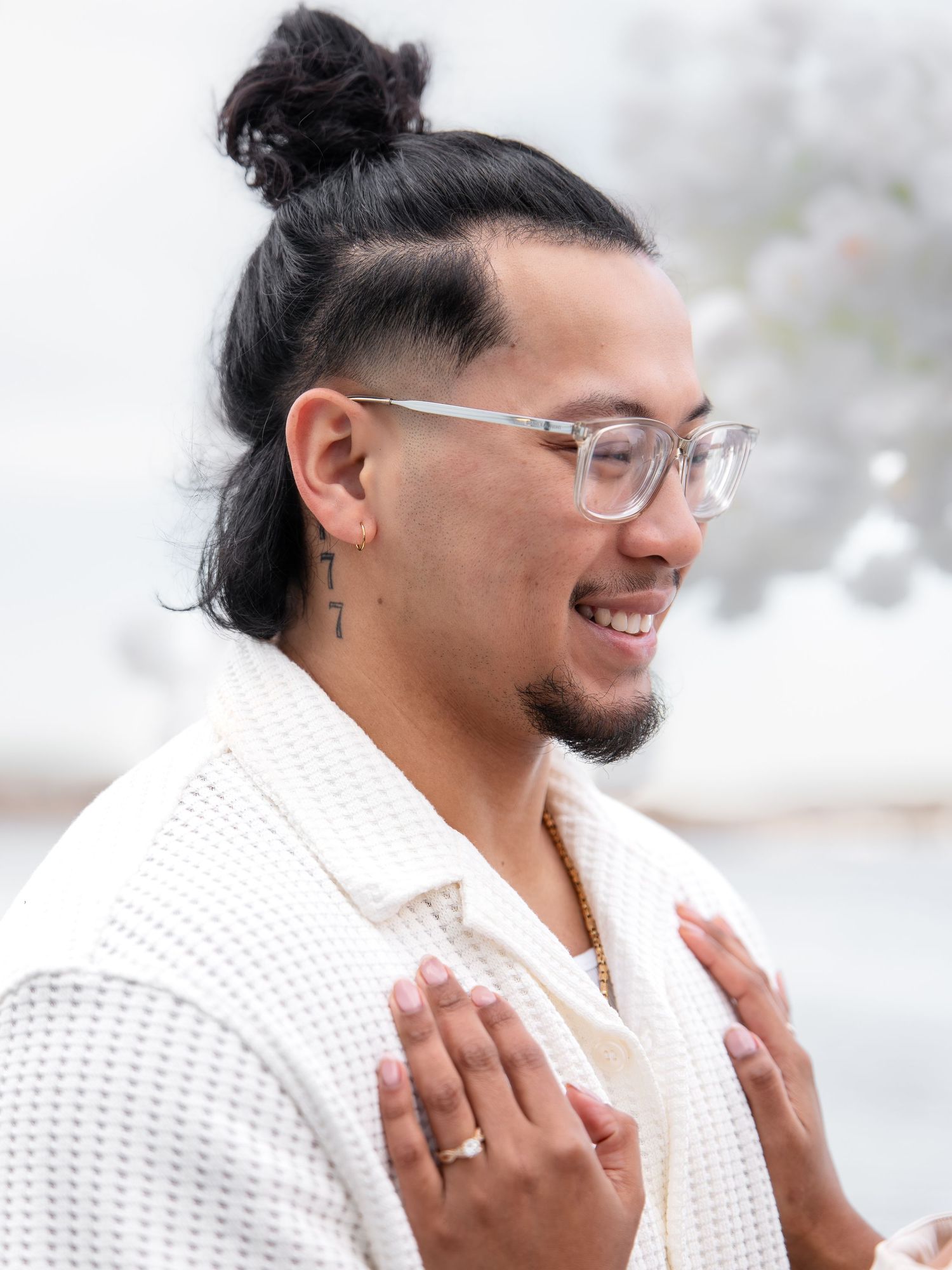 Smiling man with bun hairstyle and glasses in white waffle shirt at engagement event.