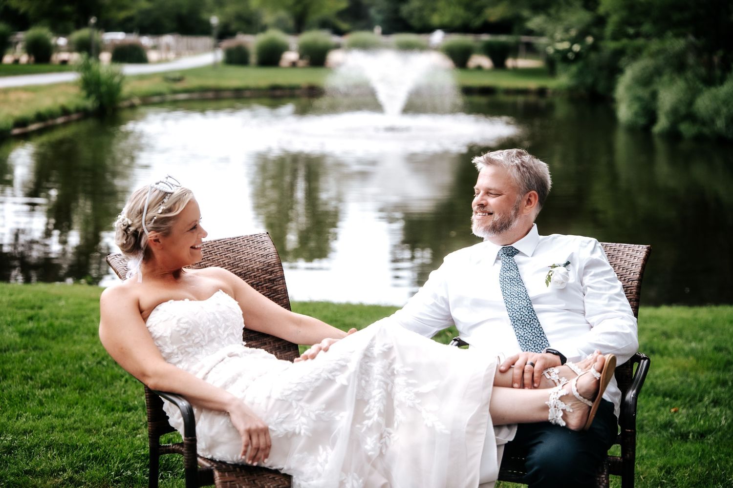 Bride and groom sitting in chairs by a pond with fountain, smiling at each other on their wedding day.