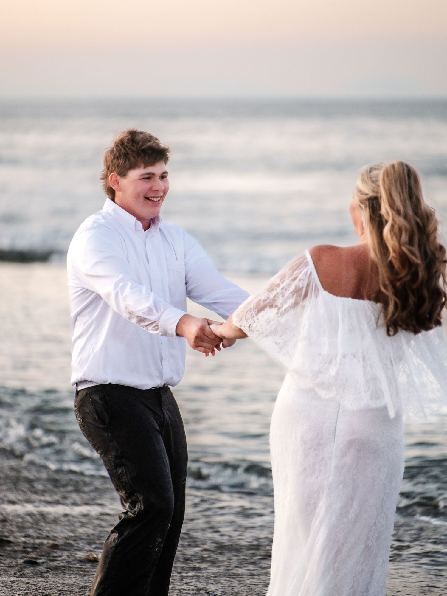 A joyful couple dances on a beach at sunset, the woman in a white lace dress and the man in a white shirt.