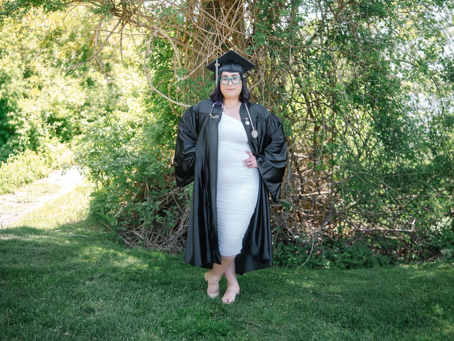 Graduate in black cap and gown standing barefoot on green grass with trees in background.