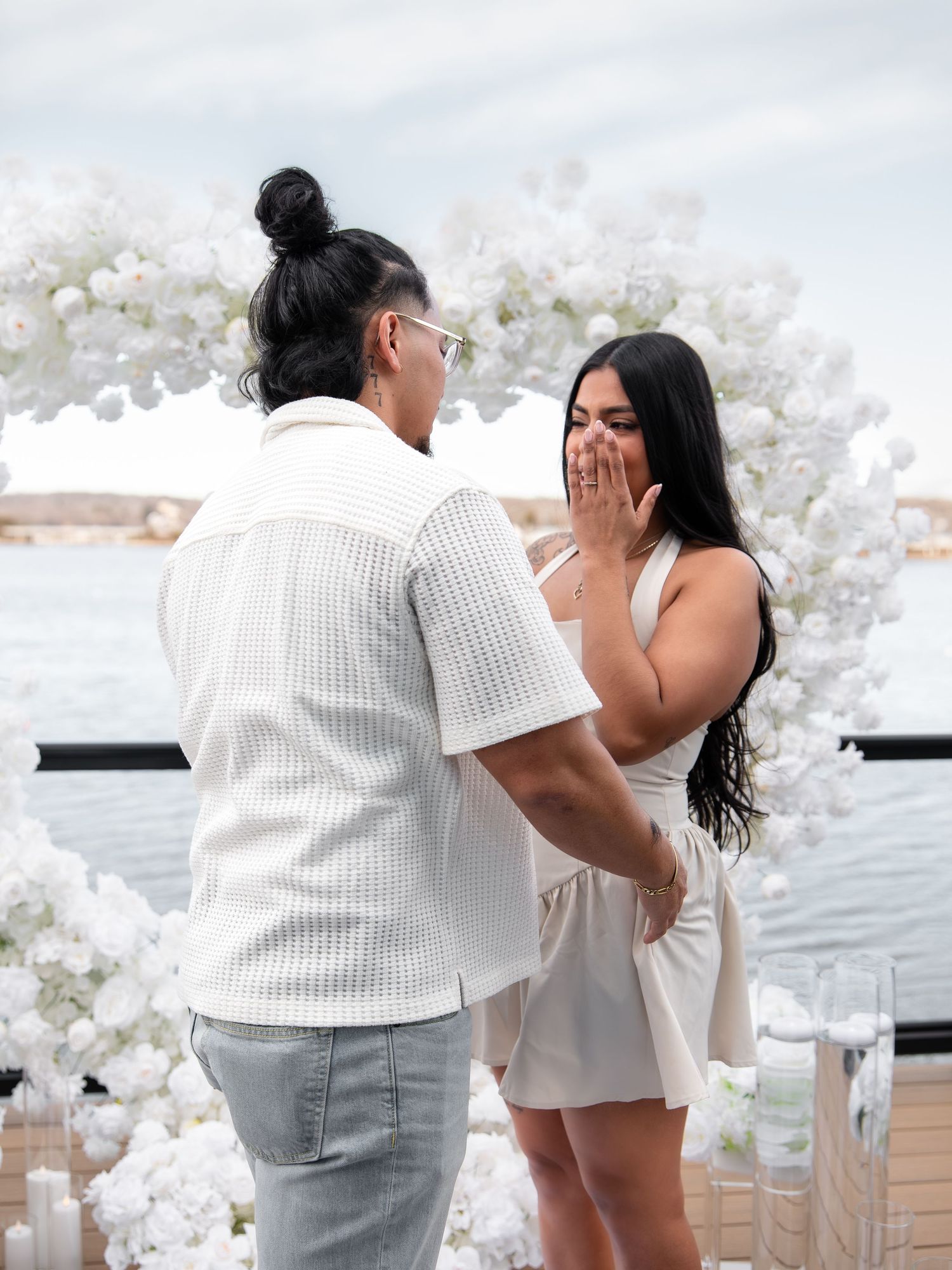 Man proposing to surprised woman near white floral arch by the water, woman covering mouth in shock.