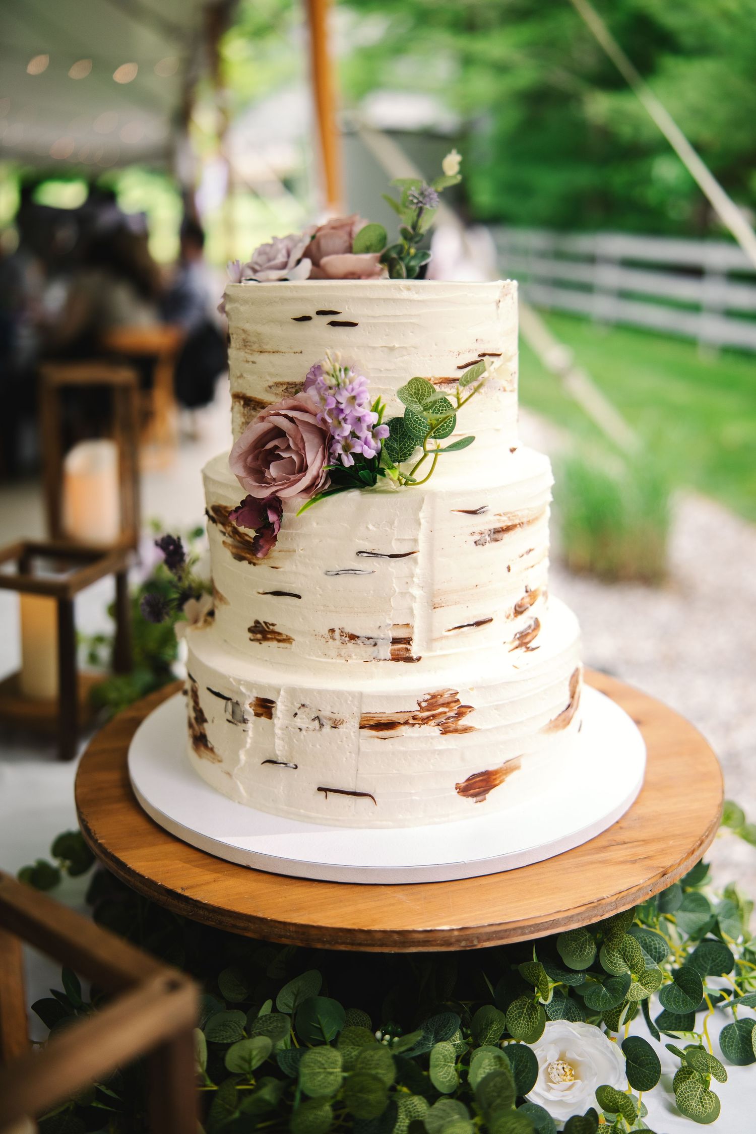 Three-tier naked wedding cake with purple roses and greenery on rustic wood stand.
