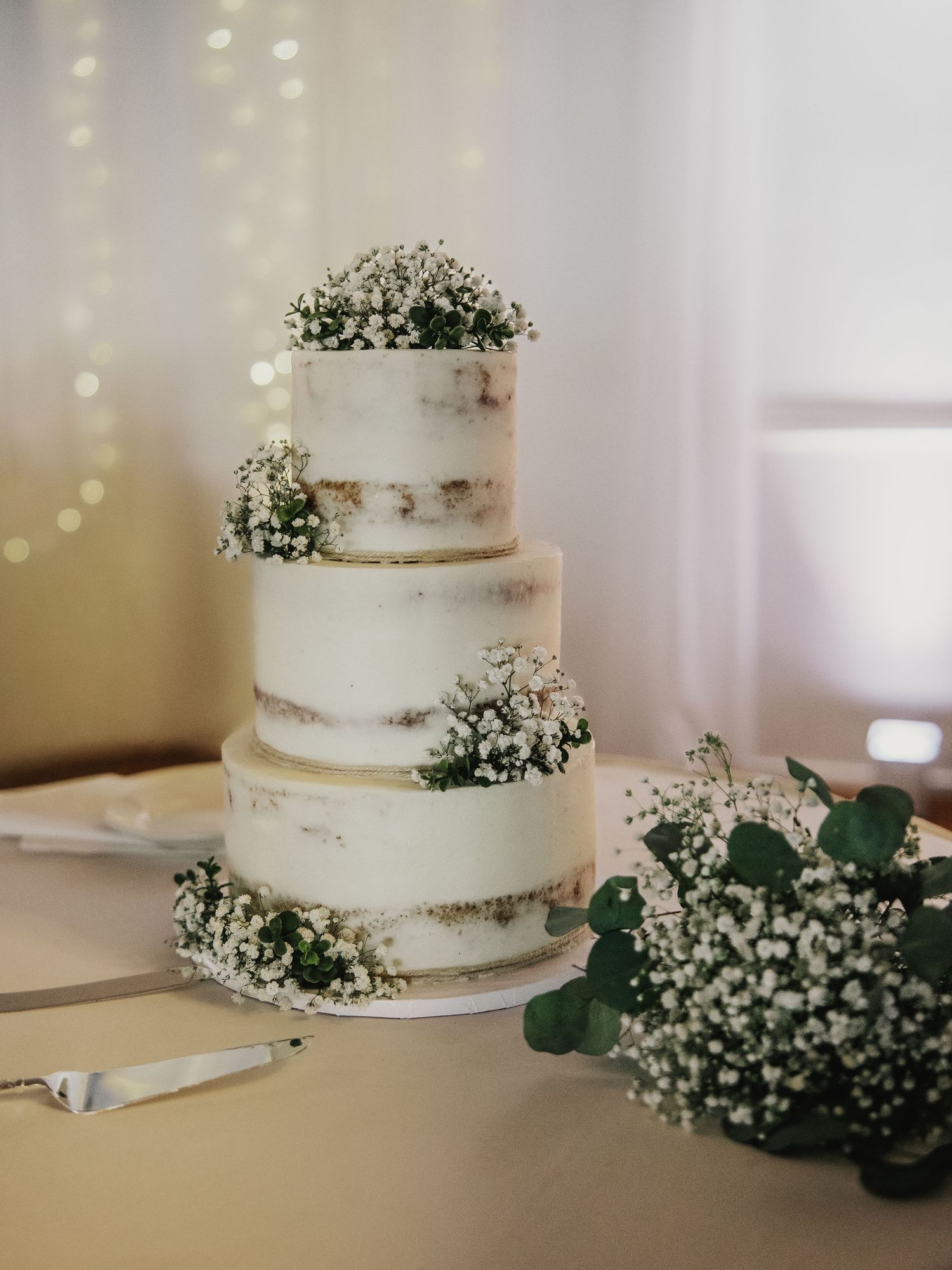 Three-tier semi-naked wedding cake decorated with baby's breath flowers on a white table with bokeh lights.