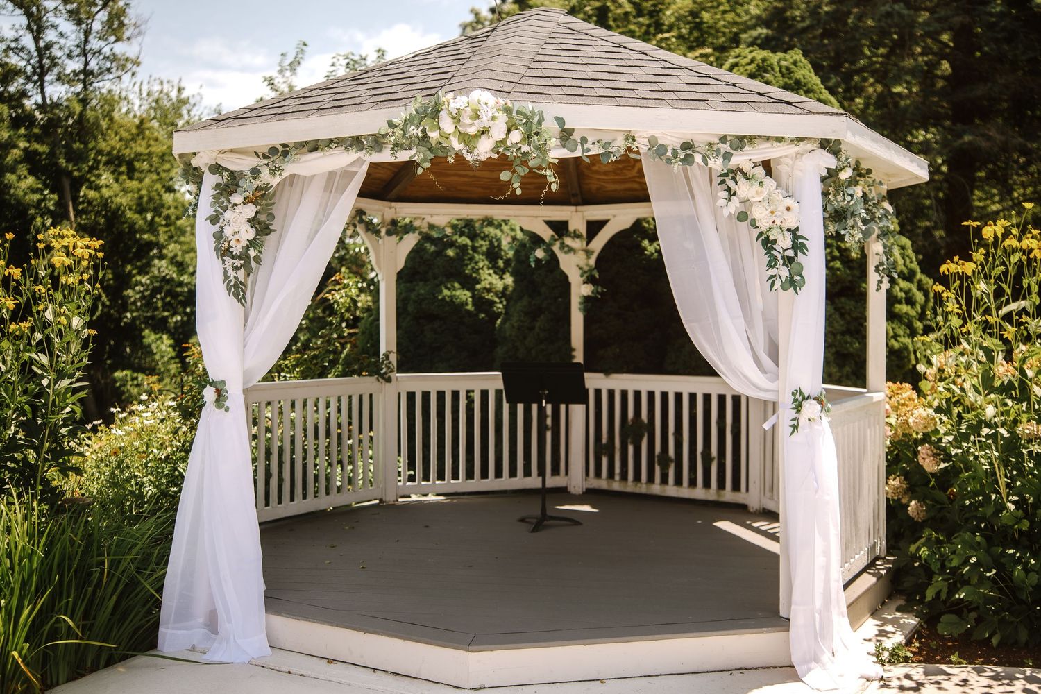 White gazebo decorated with floral arrangements and draped curtains for an outdoor wedding ceremony.