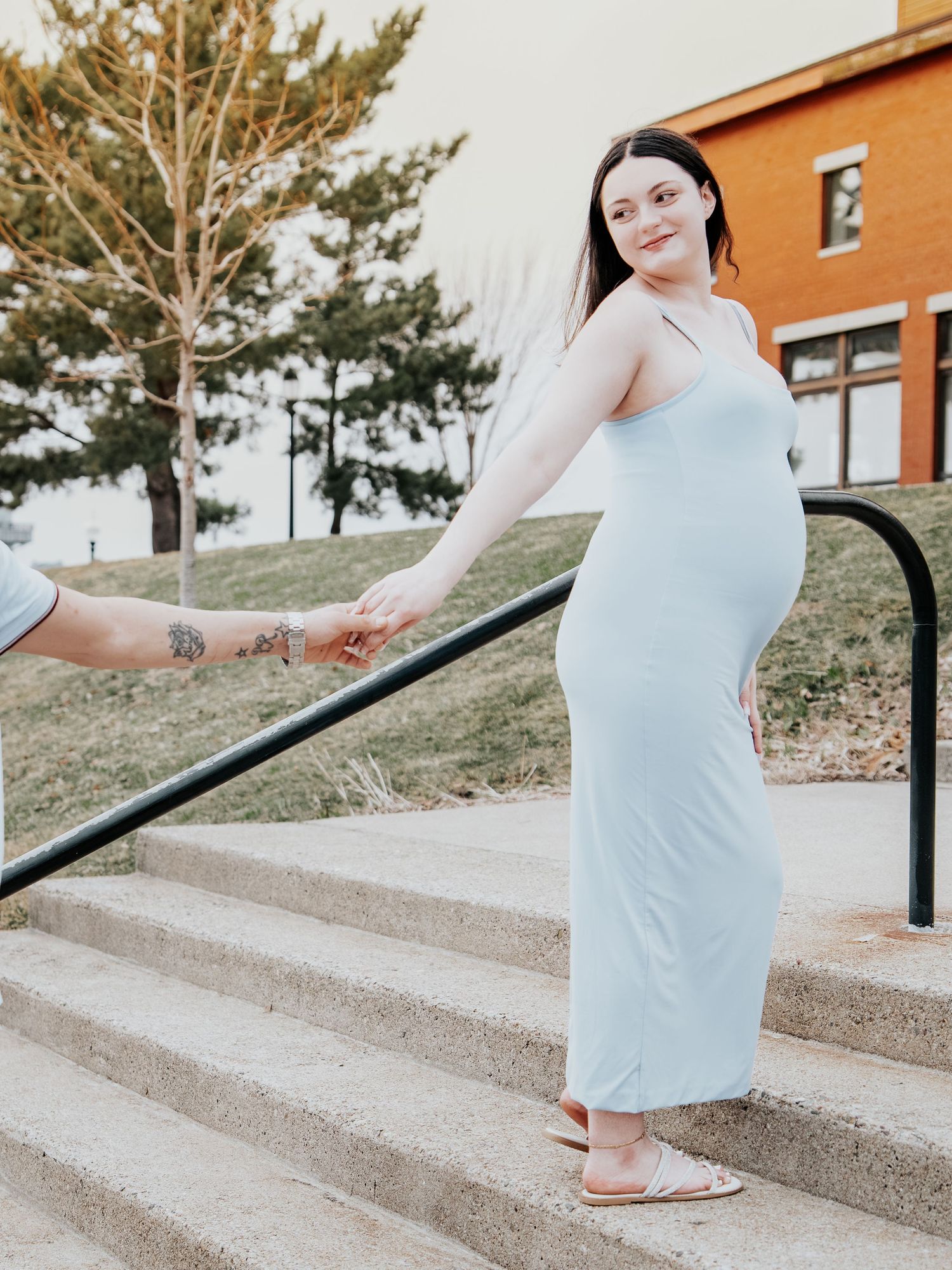 Pregnant woman in light blue maxi dress holds hands on outdoor steps during maternity photo session.