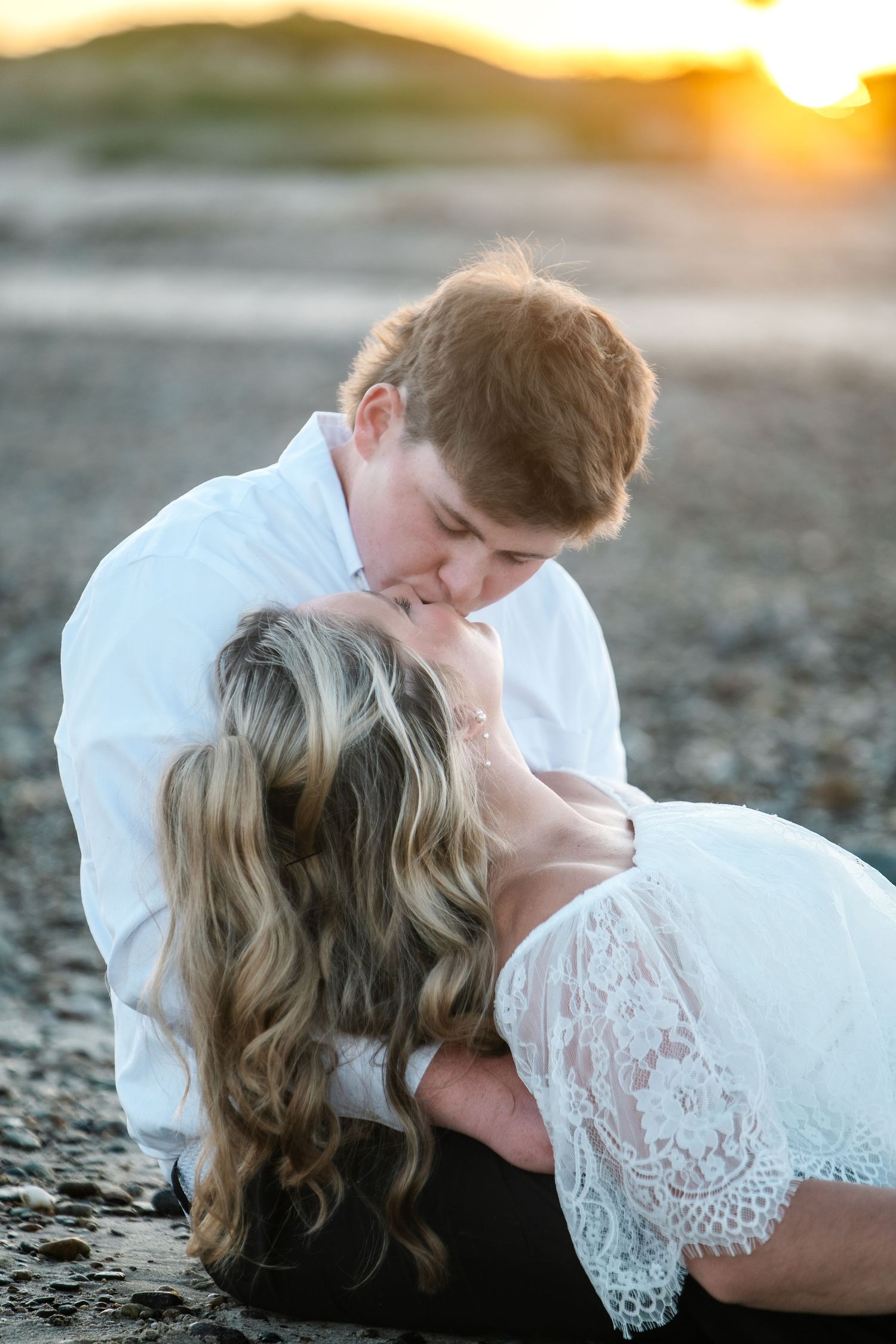 Romantic beach couple kissing at sunset, bride in white lace dress reclined against groom in white shirt.