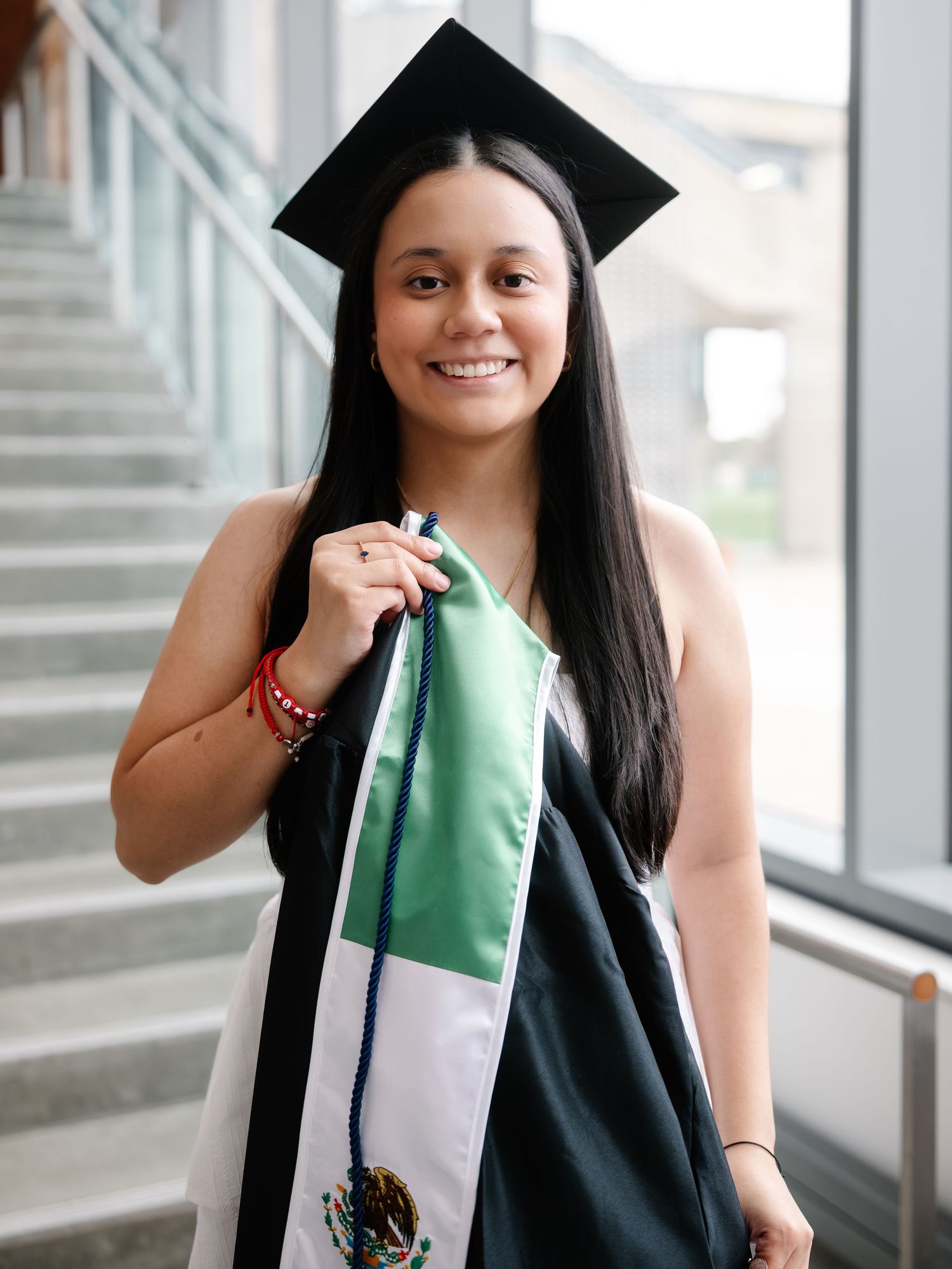 Smiling graduate in black cap holds Mexican flag stole in bright modern building interior.