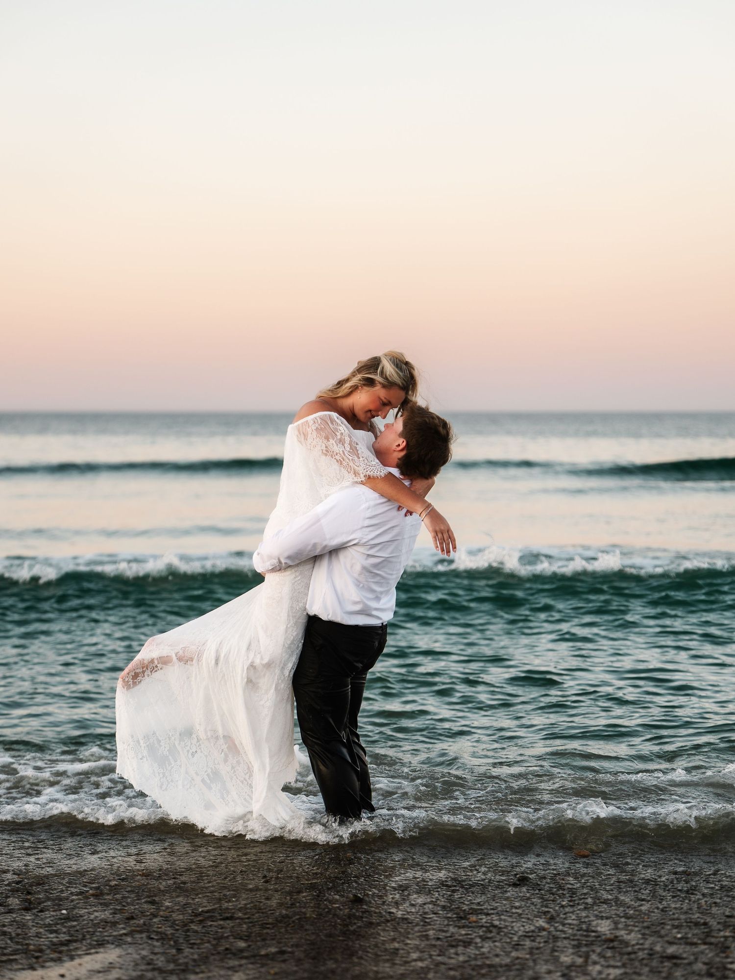Couple sharing a romantic kiss on a beach at sunset, groom lifting bride in white lace wedding gown near ocean waves.