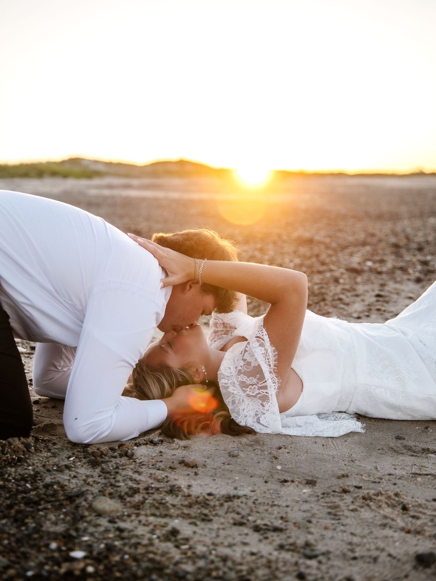Couple in wedding attire lying on beach sand sharing a kiss at golden sunset hour.
