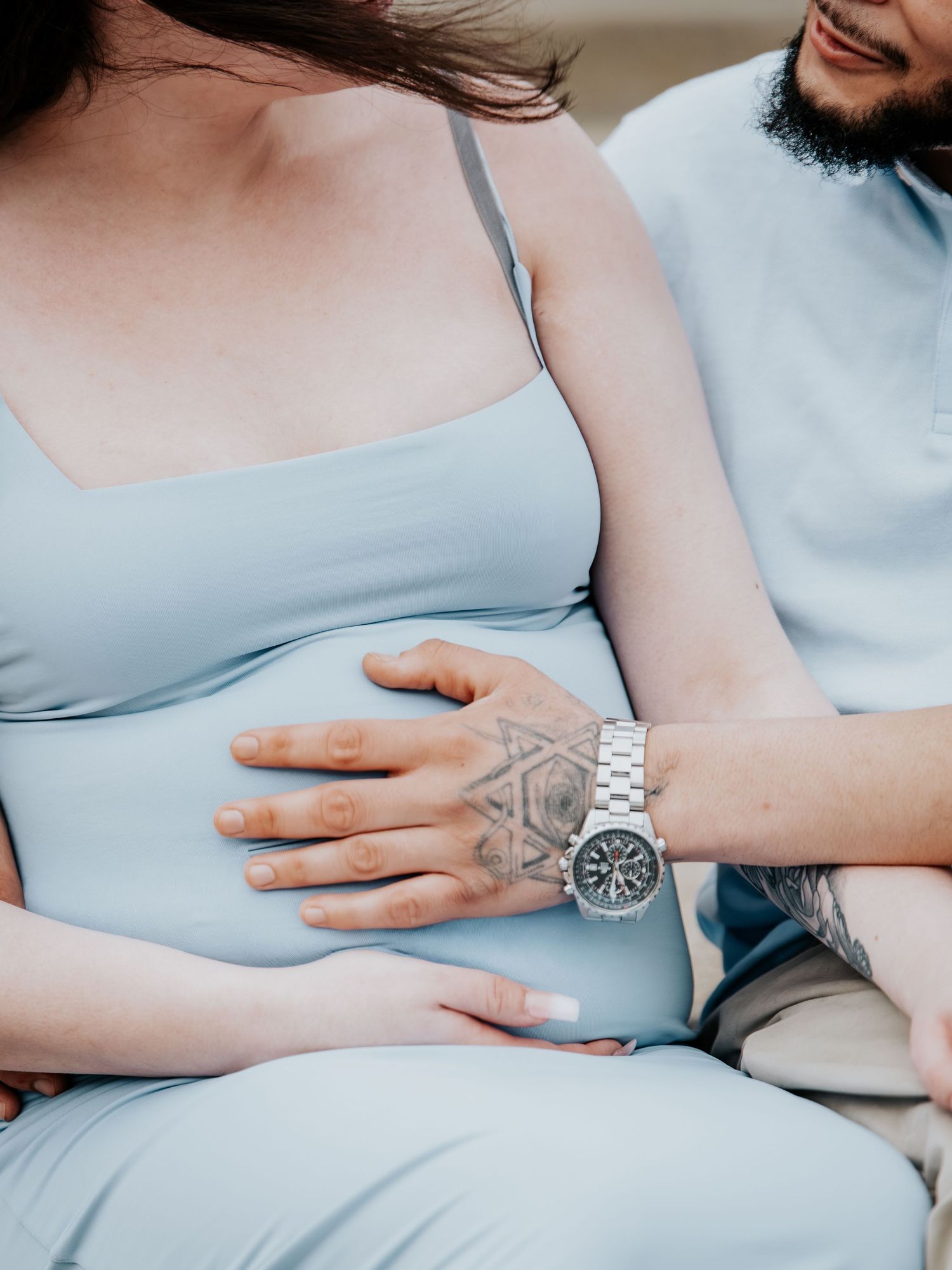 Pregnant woman in light blue dress with partner's tattooed hand and watch resting on her baby bump.