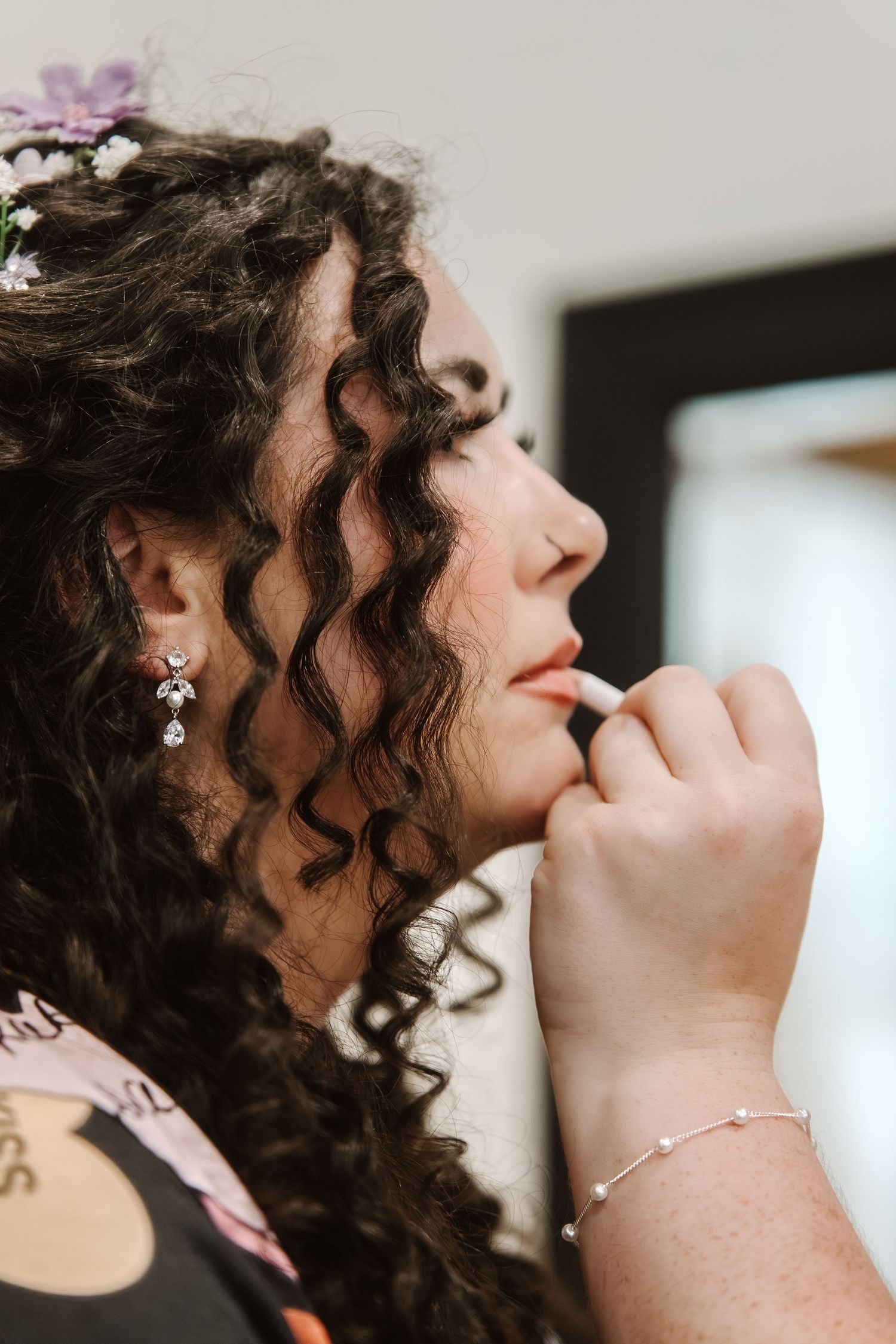 Bride with curly dark hair and floral accessories getting lipstick applied during wedding day preparations.