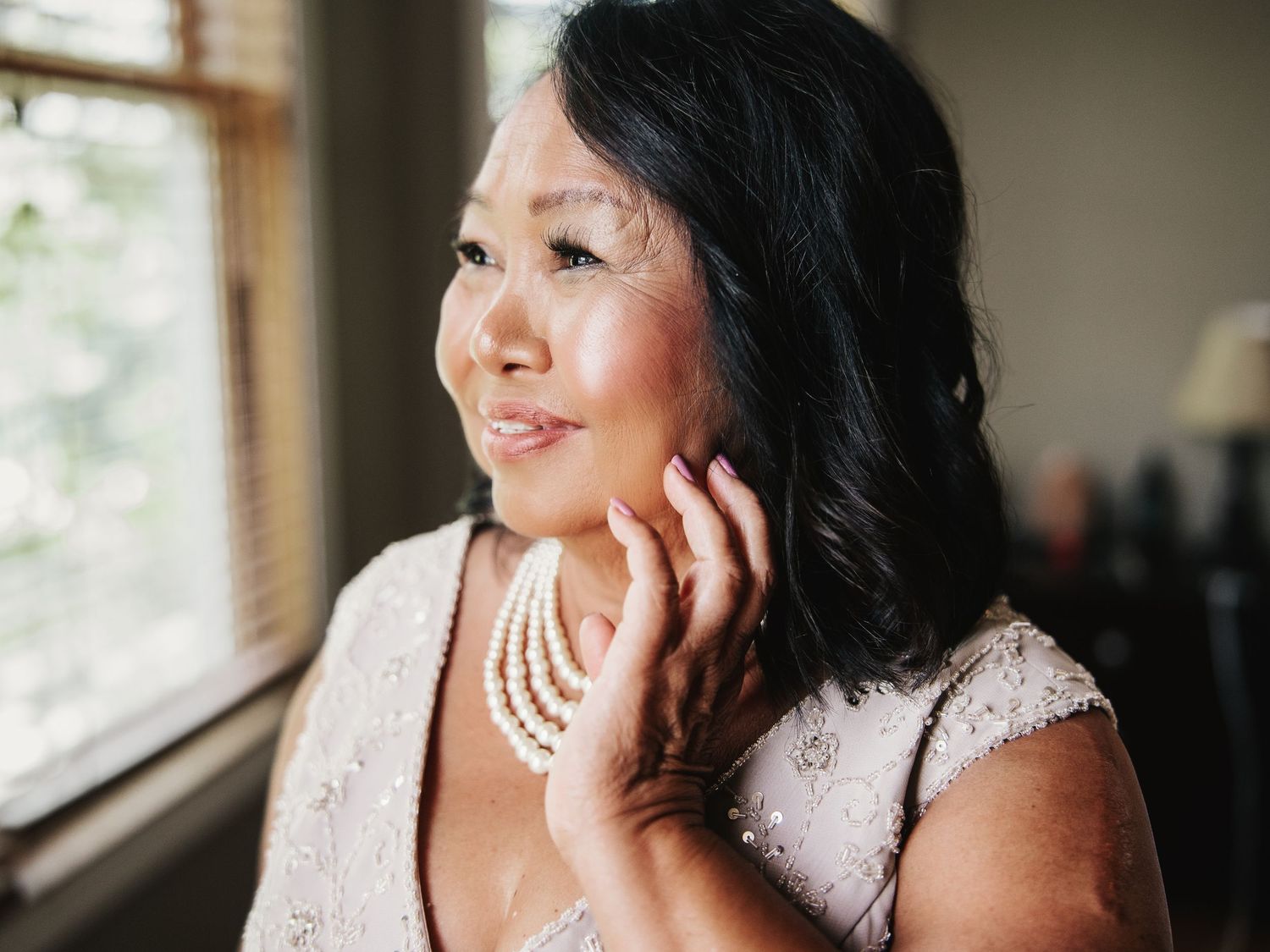 Elegant Asian woman in lace dress touching pearl necklace, smiling softly near window in natural light.