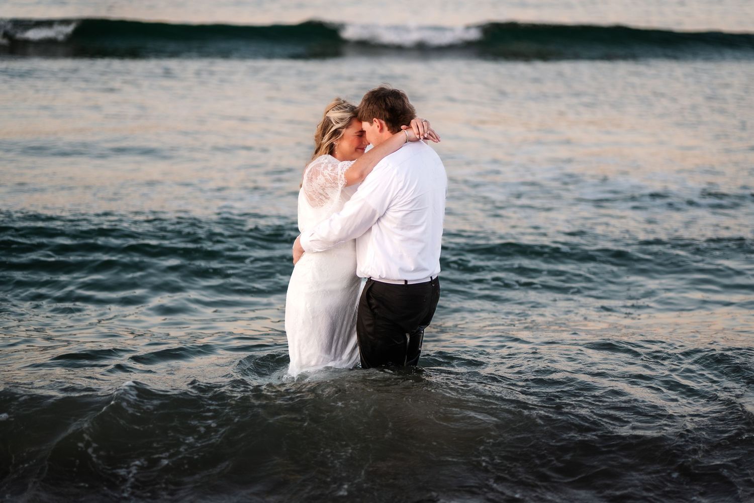 Wedding couple embracing romantically while standing in ocean water during pastel sunset beach session.