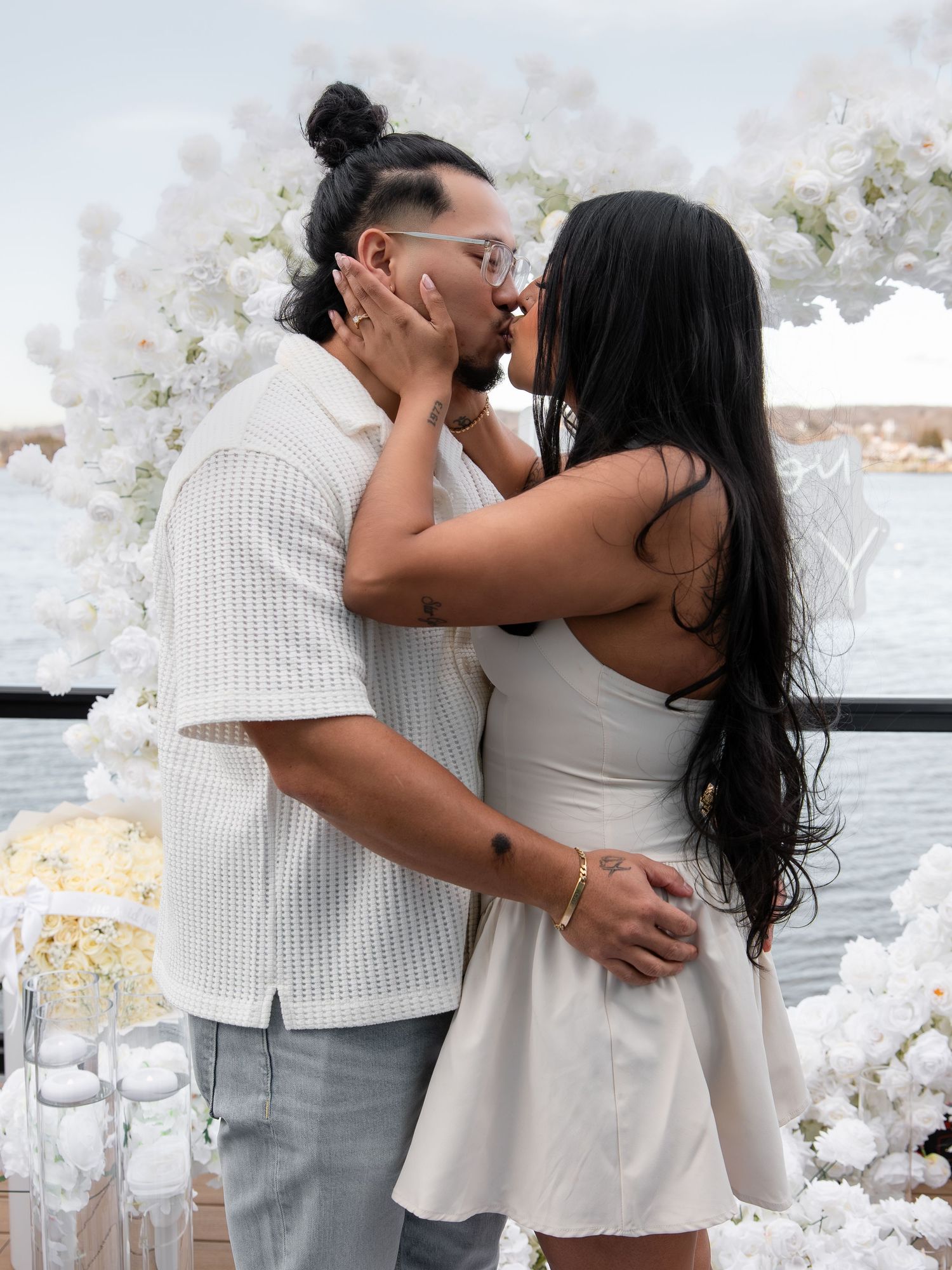 Couple sharing a romantic kiss outdoors surrounded by white floral arrangements near the water.