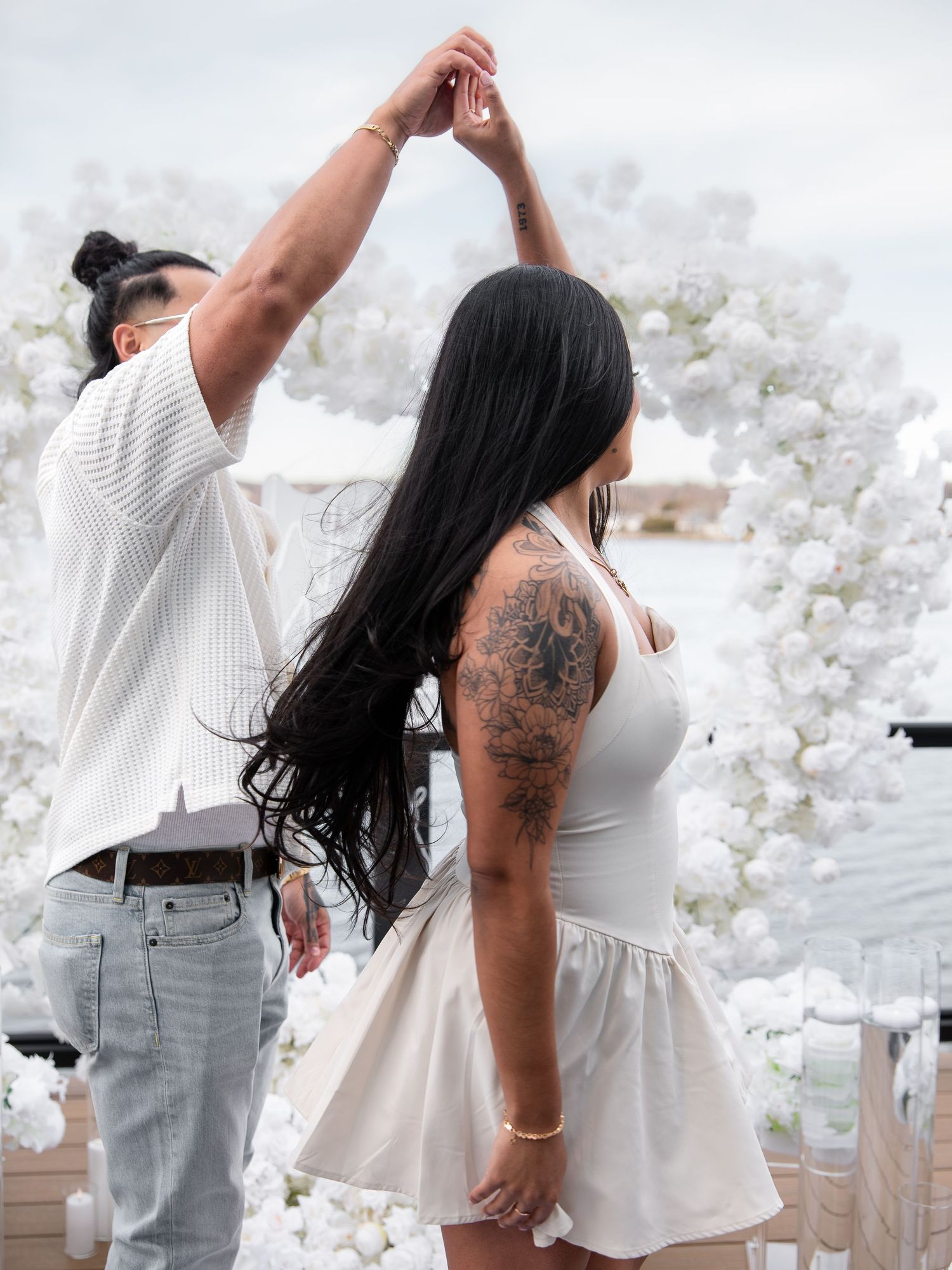 Man spinning woman in white mini dress under white floral arch near the water during proposal celebration.