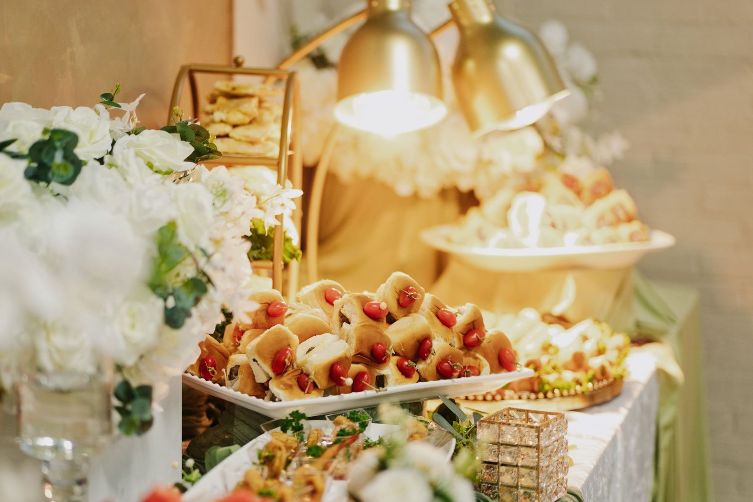 Elegant wedding buffet table with gold heat lamps illuminating mini hot dogs and fresh strawberries beside white floral arrangements.