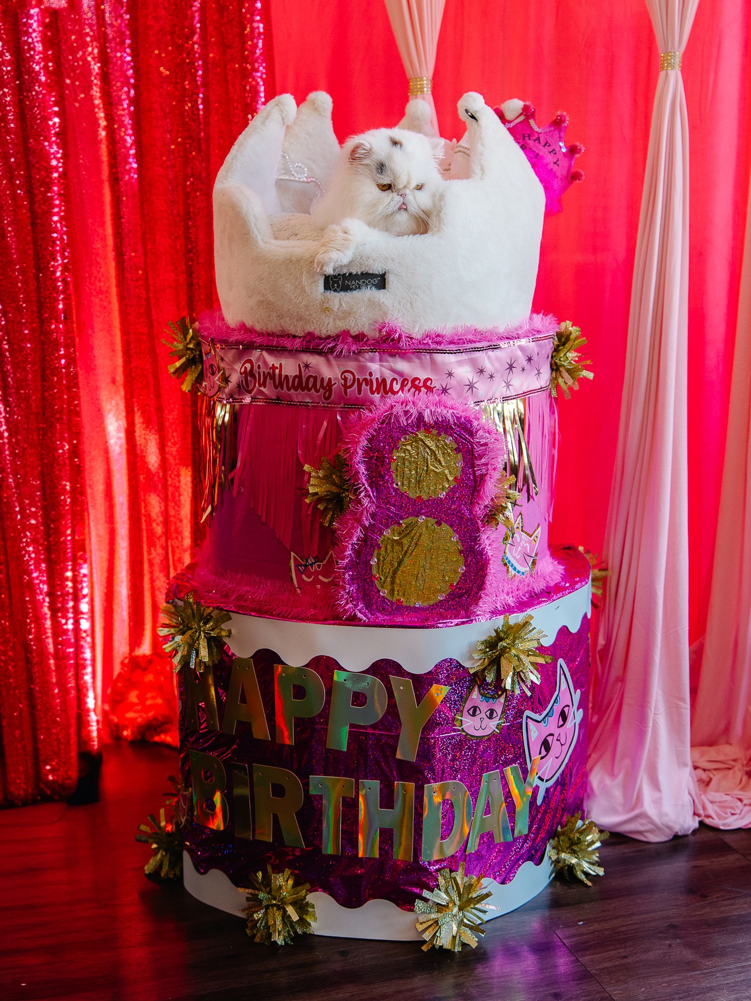 Elaborate three-tier pink and gold Happy Birthday cake decoration with a white cat sitting on top.