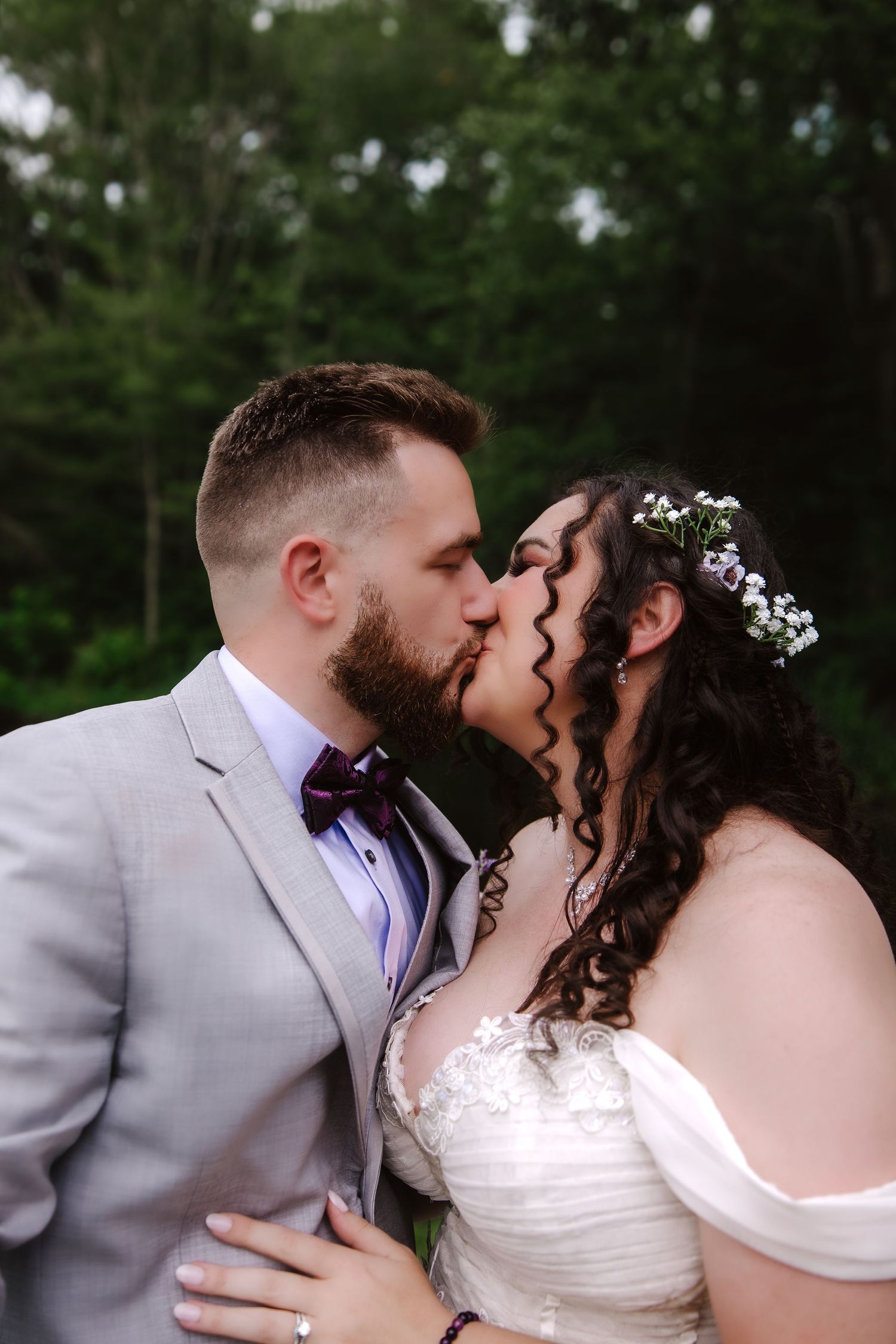 Bride and groom share a kiss outdoors, bride in off-shoulder lace gown with floral crown.