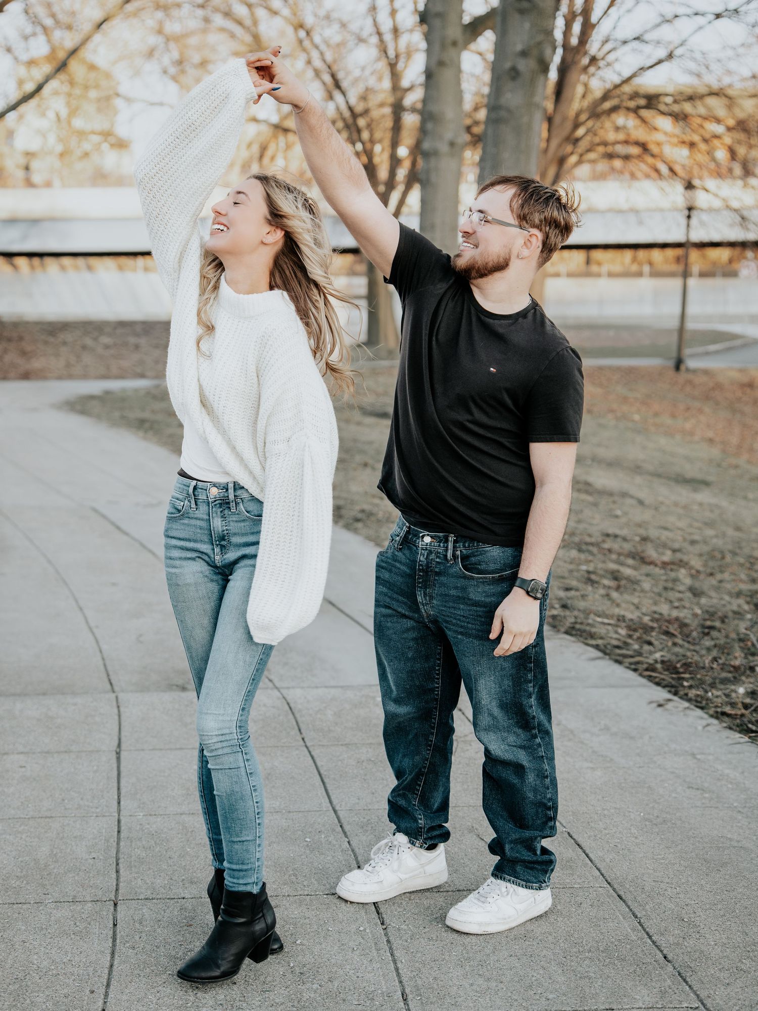 Couple dancing outdoors on a park path, woman in white sweater twirling in black boots, man in black tee smiling.