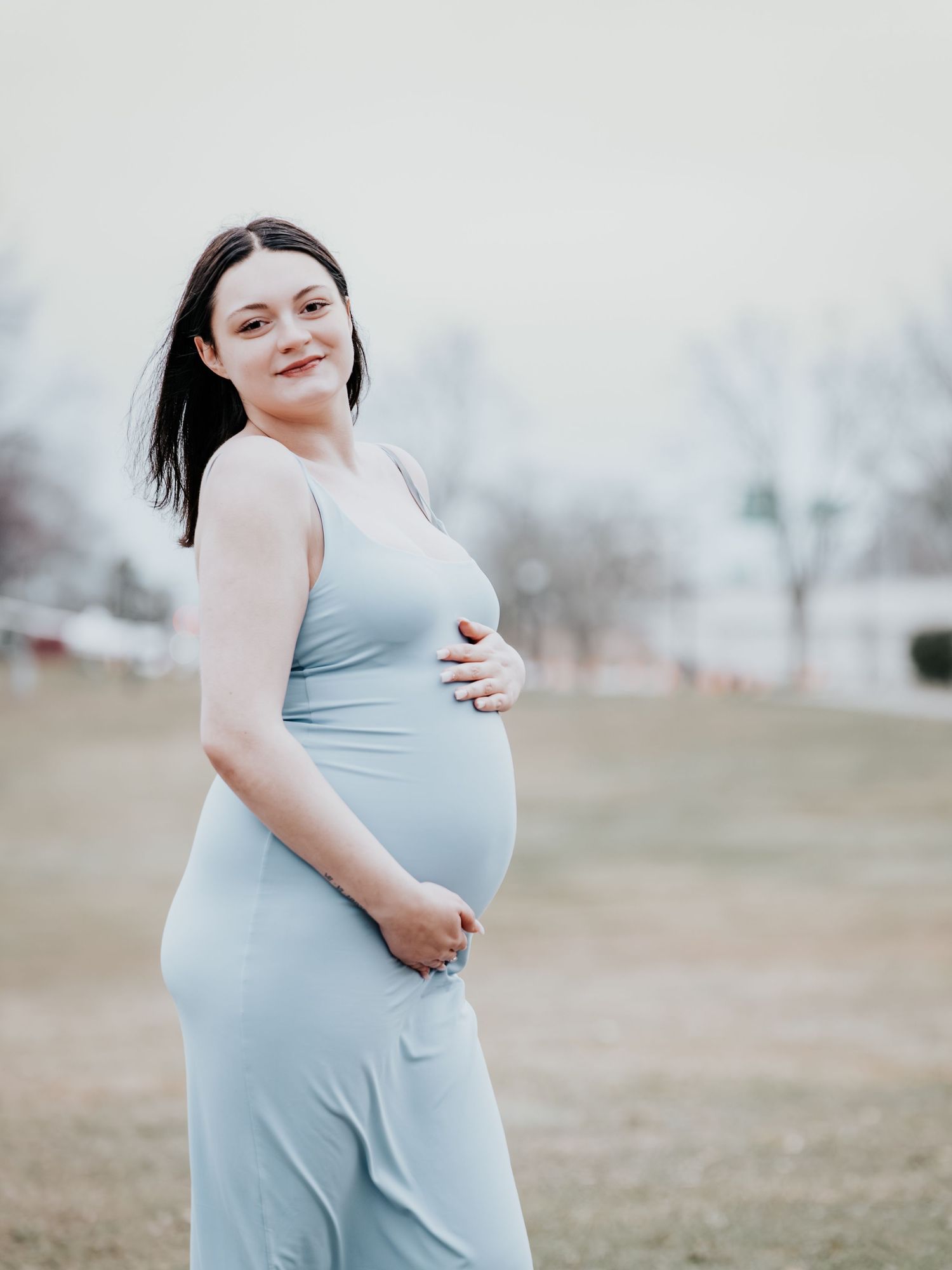 Smiling pregnant woman in a light blue maternity dress cradling her baby bump outdoors in a park.
