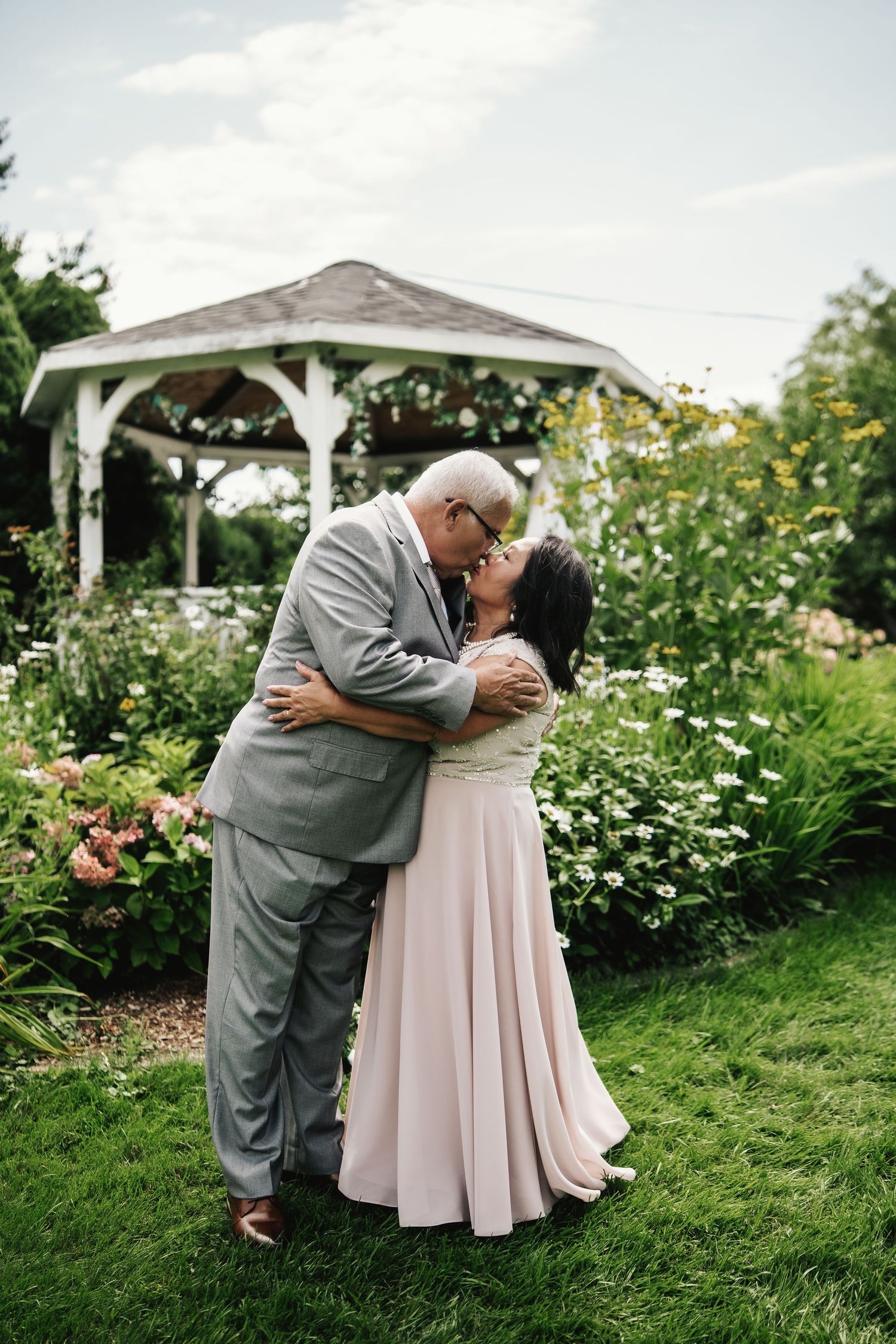Older couple sharing a romantic kiss in a garden with a white gazebo and colorful flowers in the background.