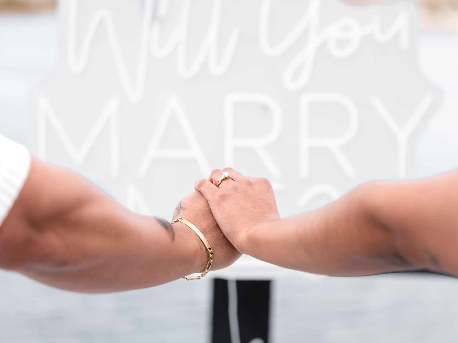 Two people holding hands with a gold bracelet and ring, in front of a Will You Marry Me sign.