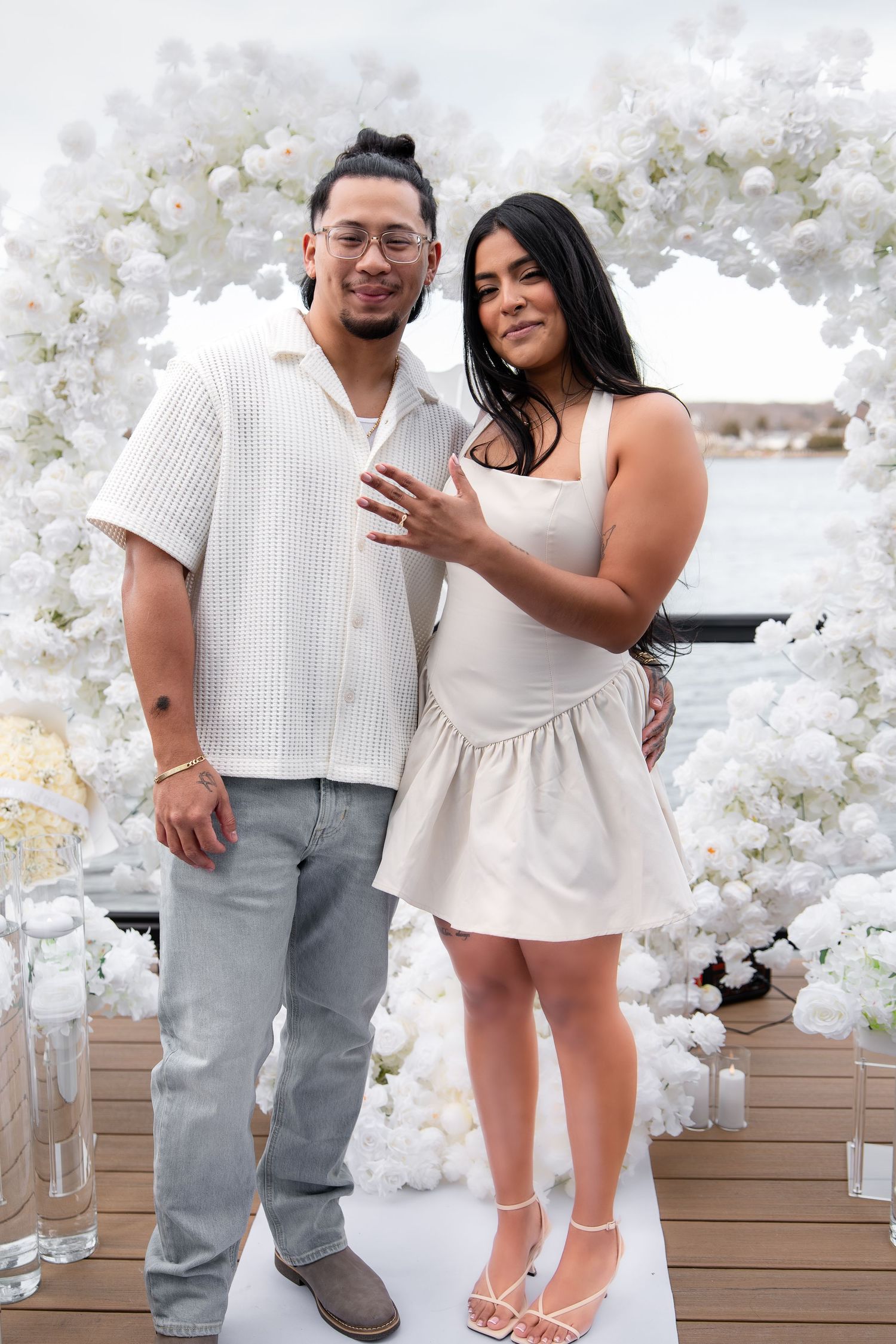 A couple poses together in front of a white floral arch on a waterfront deck, dressed in coordinating white outfits.