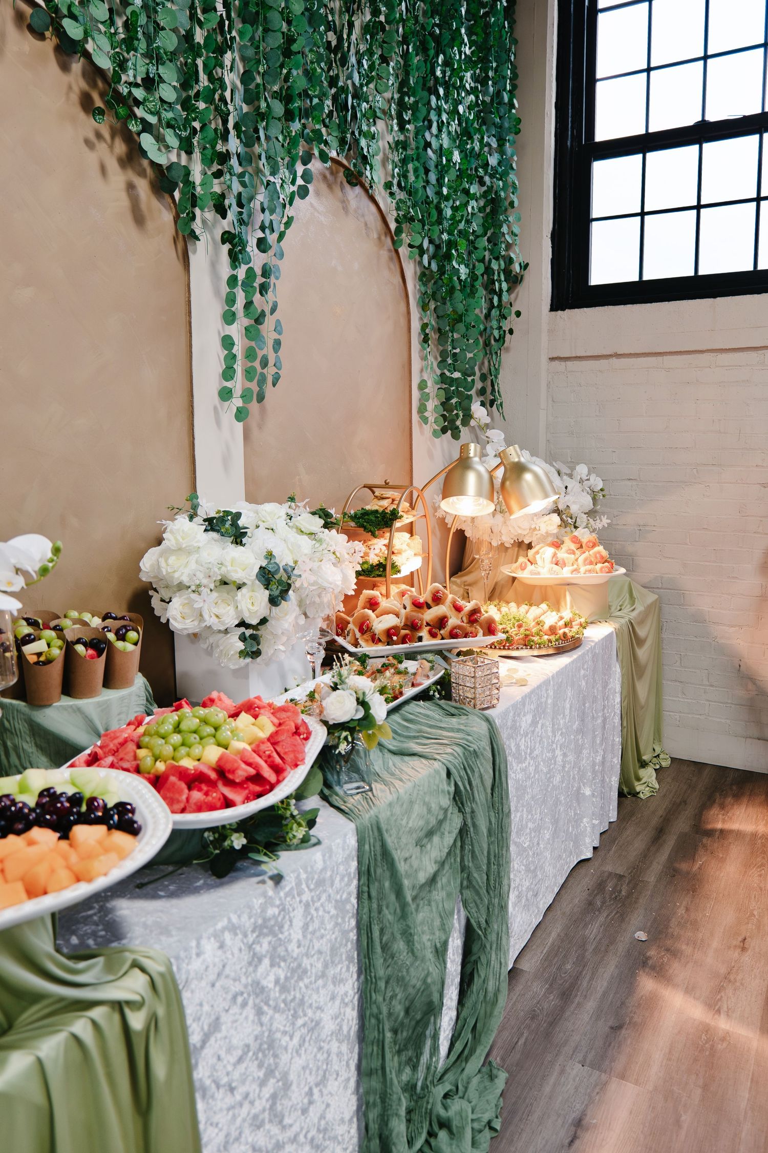 Wedding reception food table featuring fresh fruit platter, white floral centerpieces, sage green draping, and hanging greenery backdrop.