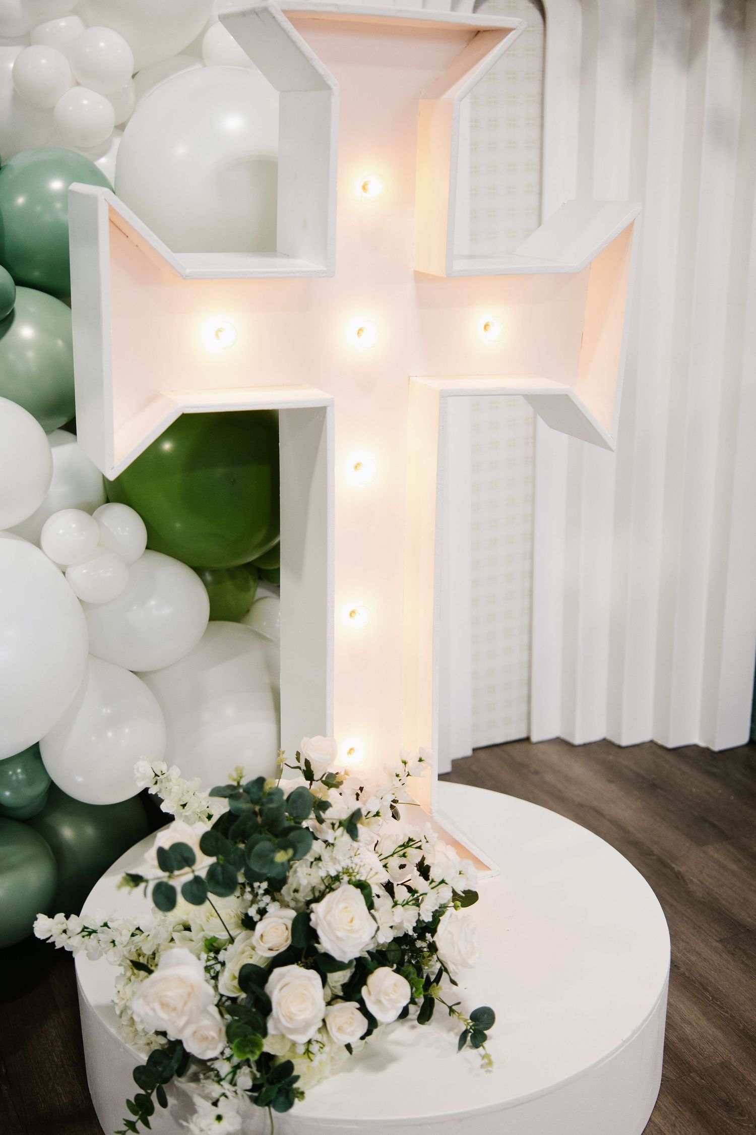 Illuminated white cross marquee light surrounded by sage green and white balloon garland with roses.