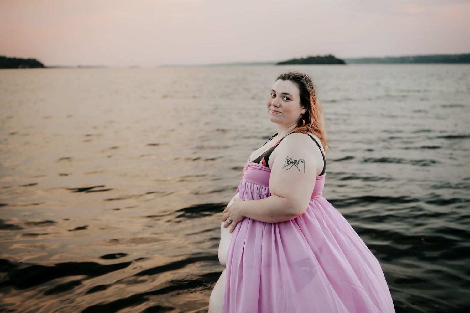 Woman in pink gown standing by lake at sunset with tattoo on arm.