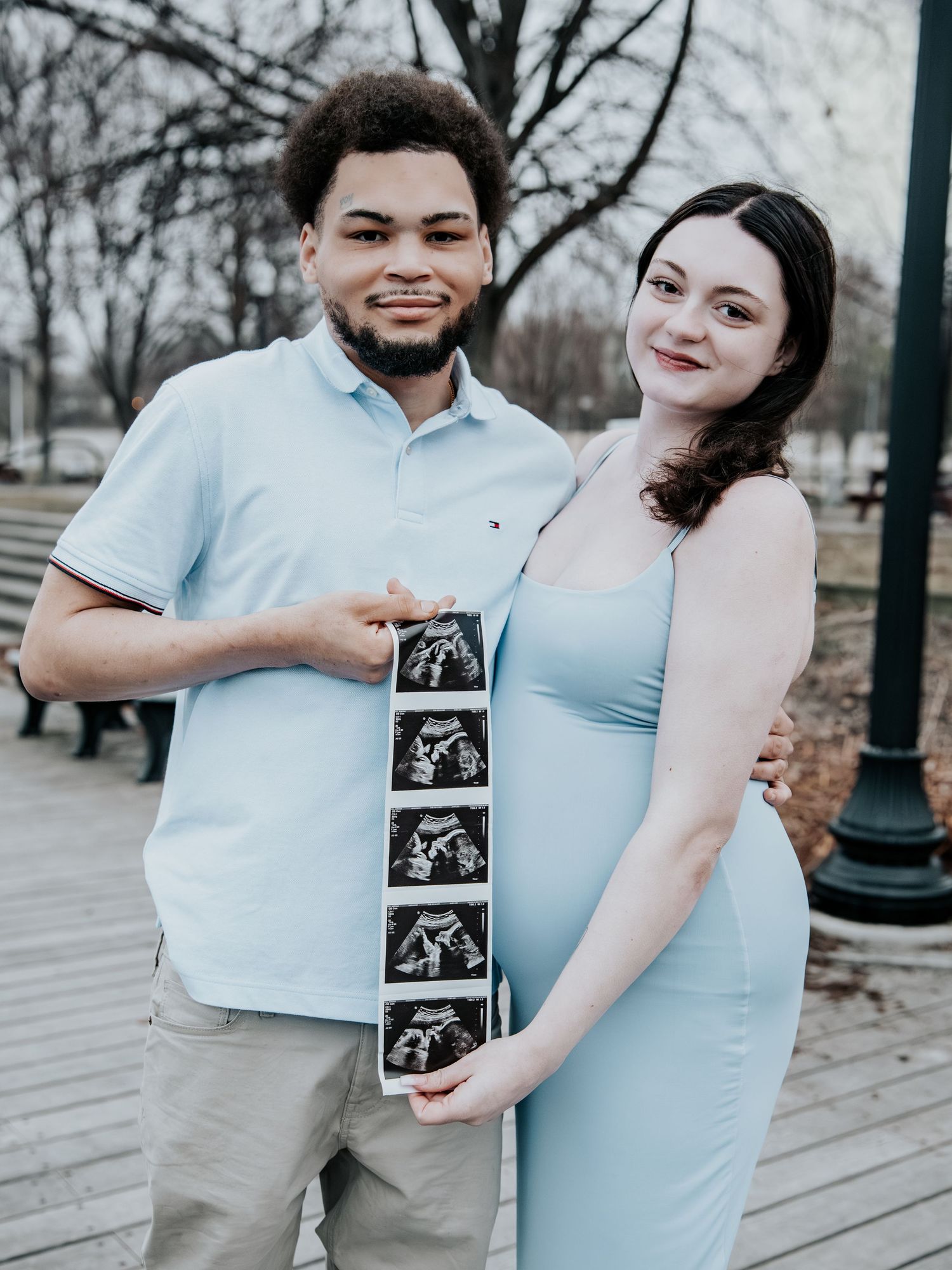 Expecting couple in matching blue outfits holds ultrasound photos during outdoor maternity photo session in park.