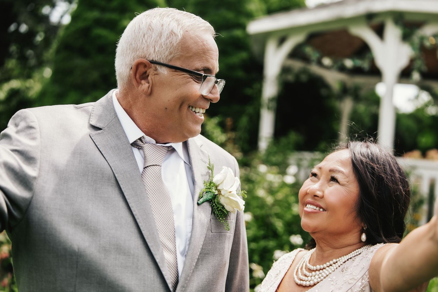 Smiling older couple in wedding attire sharing a joyful moment outdoors near a white gazebo.