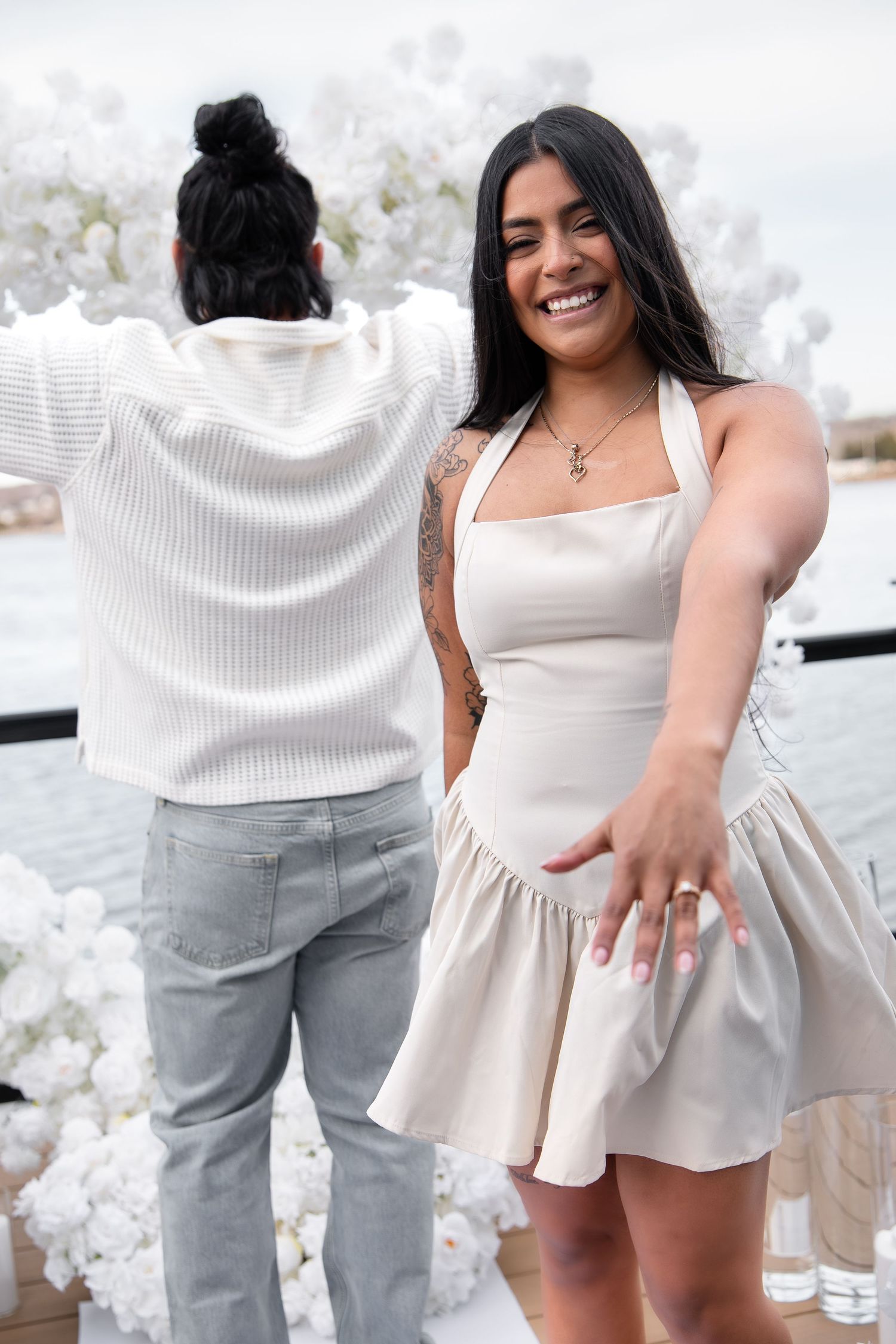 Smiling woman in cream mini dress showing off engagement ring while partner poses by white floral arch.