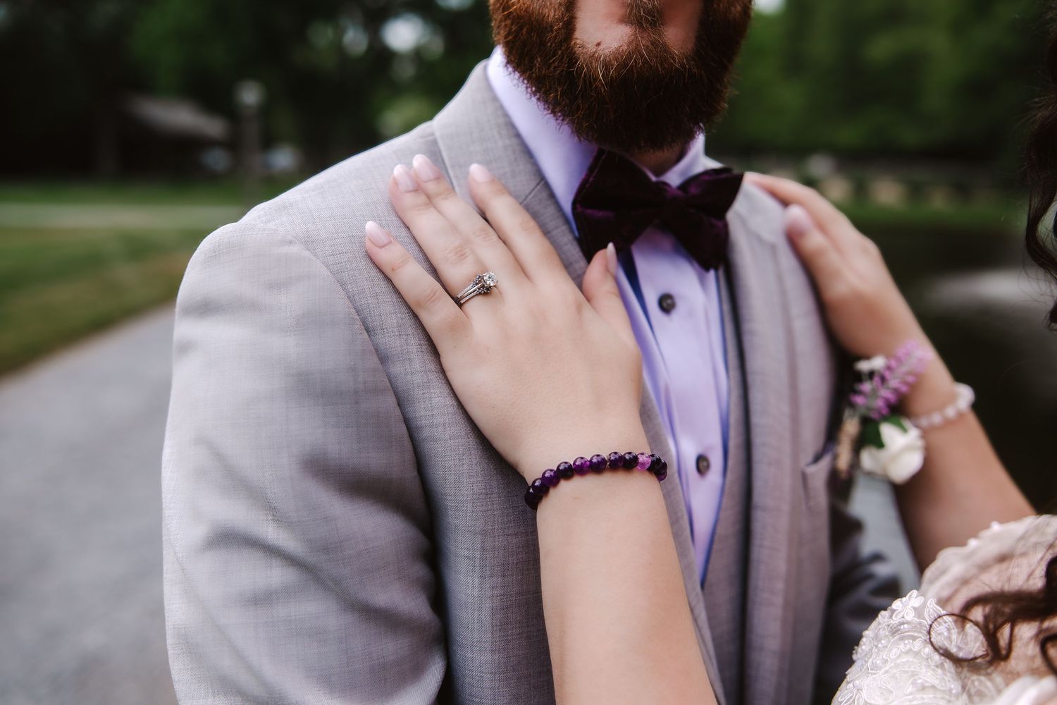 Bride places hand on groom's gray suit, showcasing engagement ring and purple beaded bracelet.